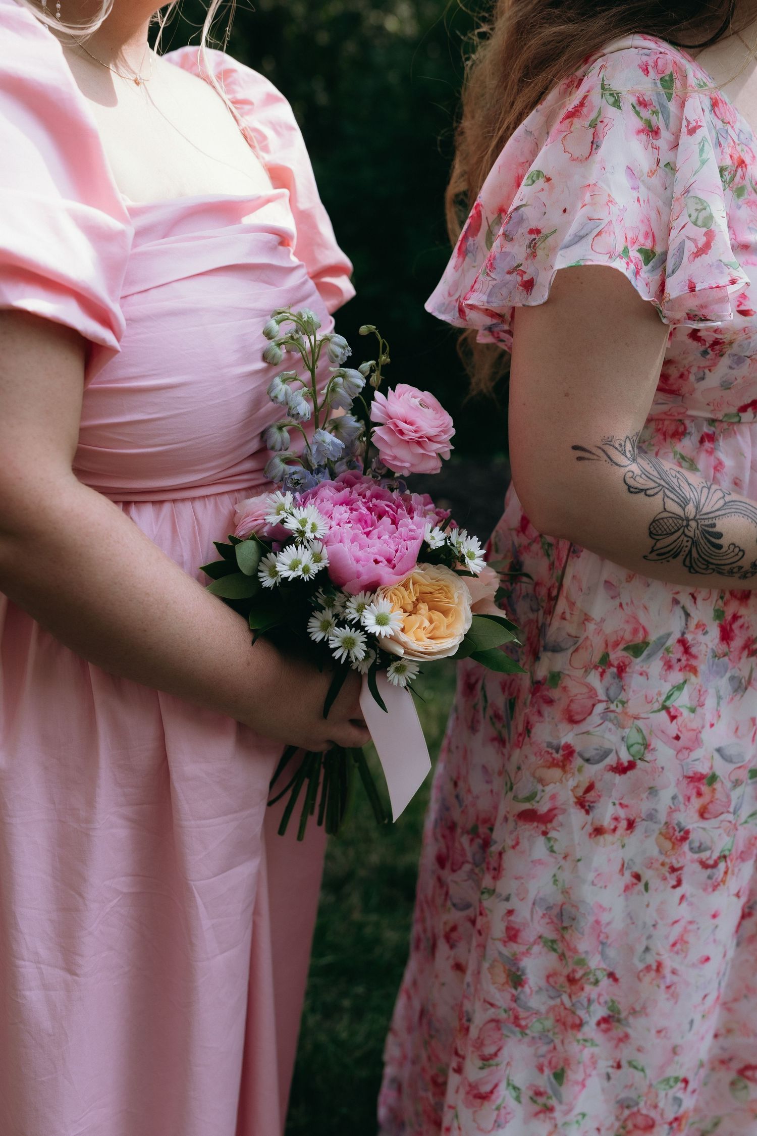 Two women in pink floral dresses hold a colorful bouquet of peonies and wildflowers at a wedding.