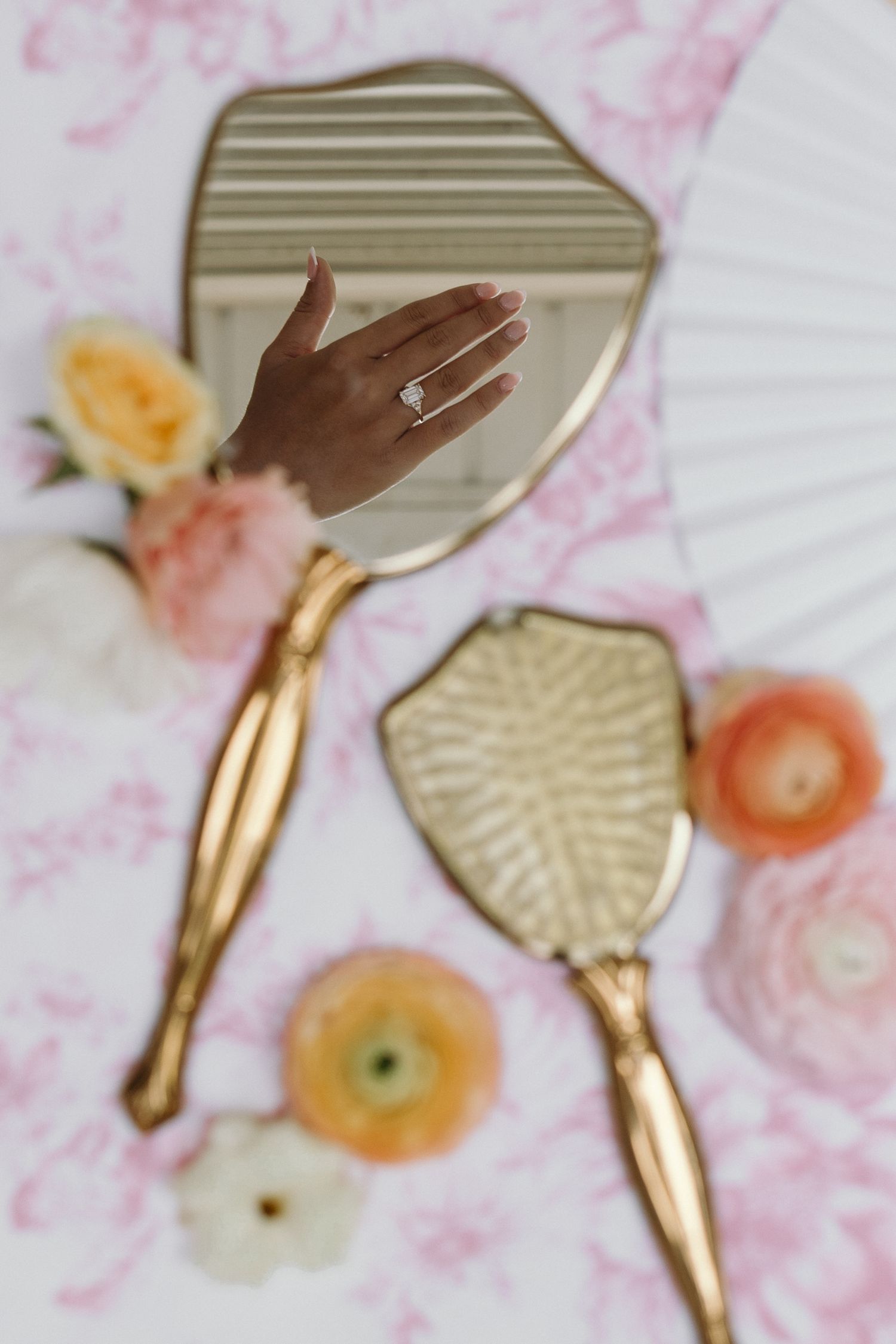 Hand with diamond engagement ring reflected in vintage gold hand mirror, surrounded by pastel flowers on pink floral background.
