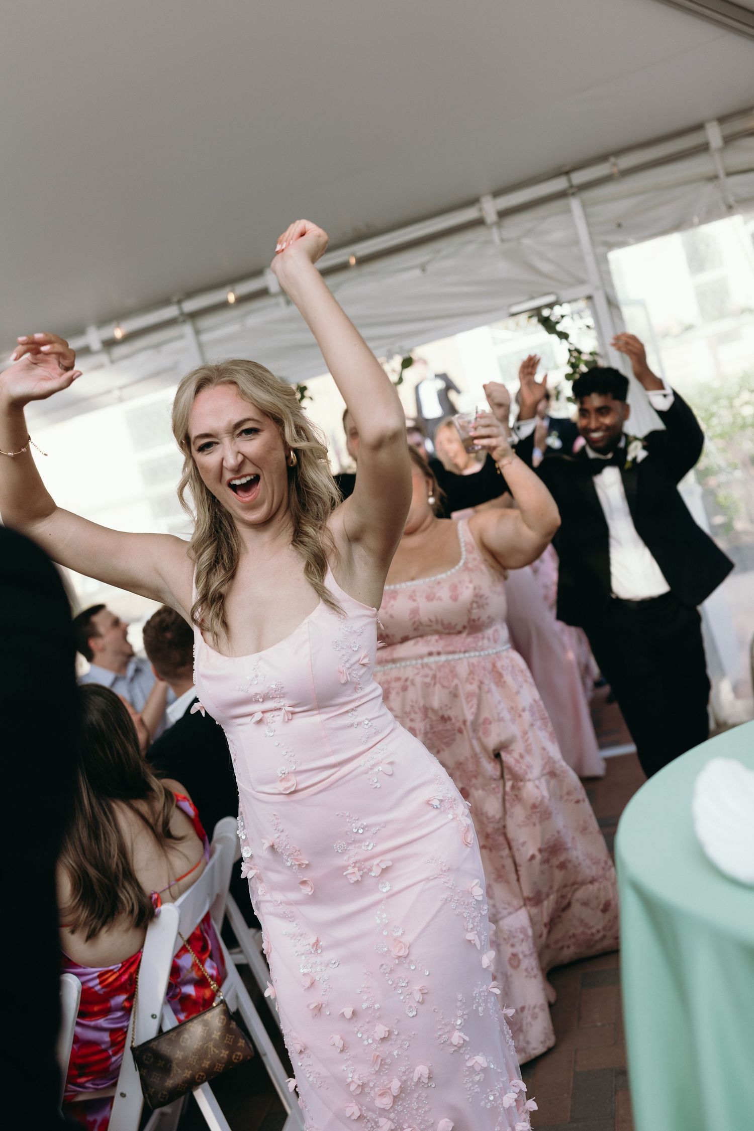 A joyful woman in a pink floral dress dances with arms raised at an elegant wedding reception under a white tent.
