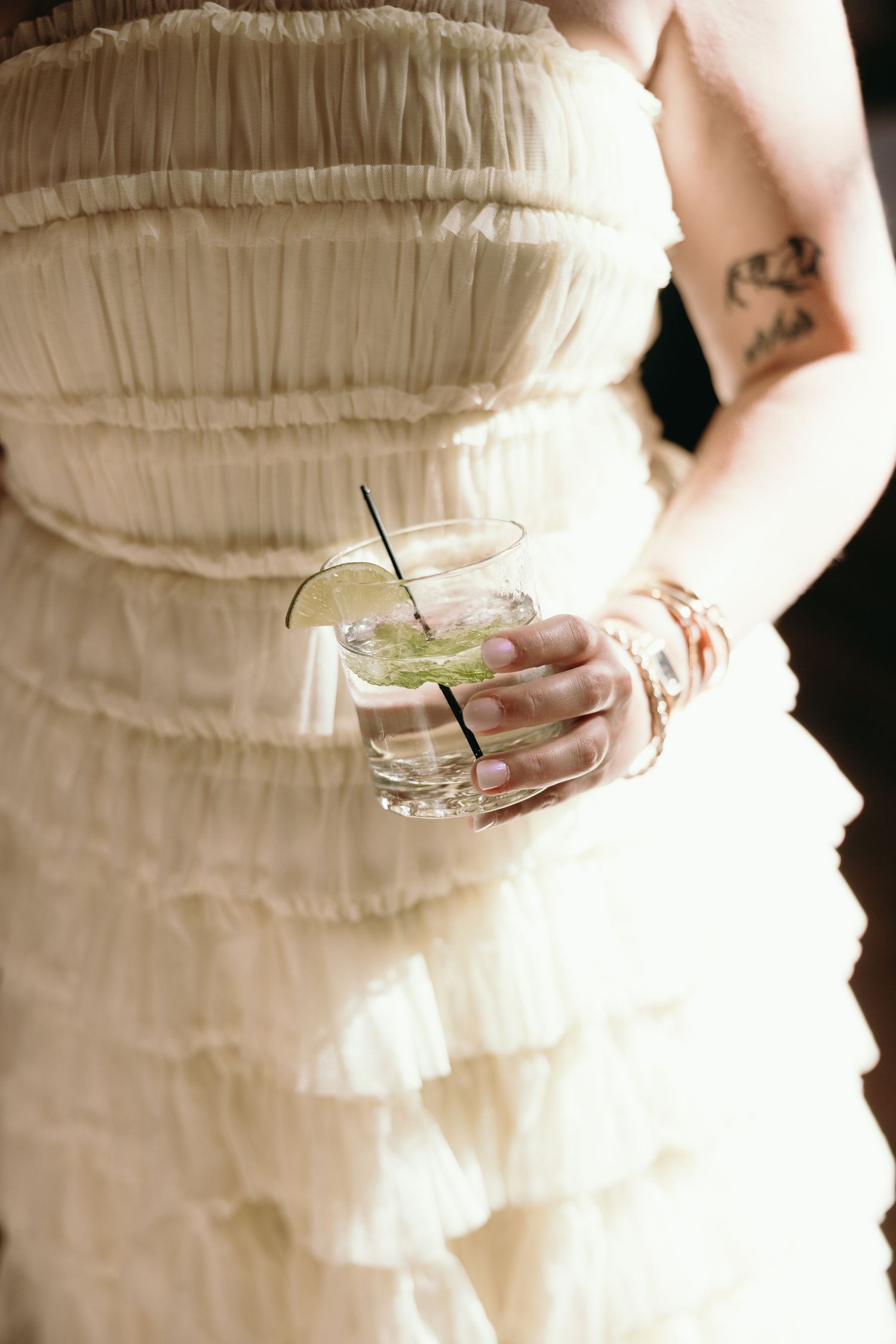 Someone in a layered cream-colored dress holds a cocktail glass with lime garnish and clear liquid.