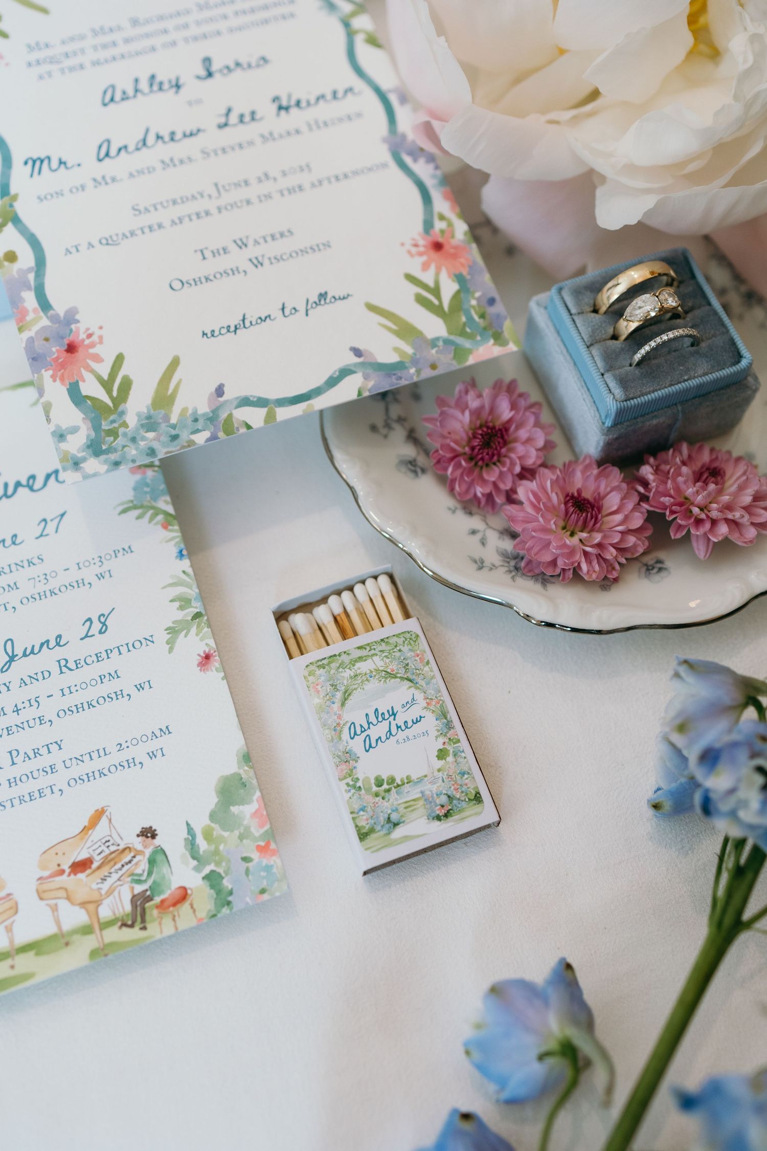 Wedding invitation with floral design displayed alongside rings, flowers, and customized matchbox on white surface.