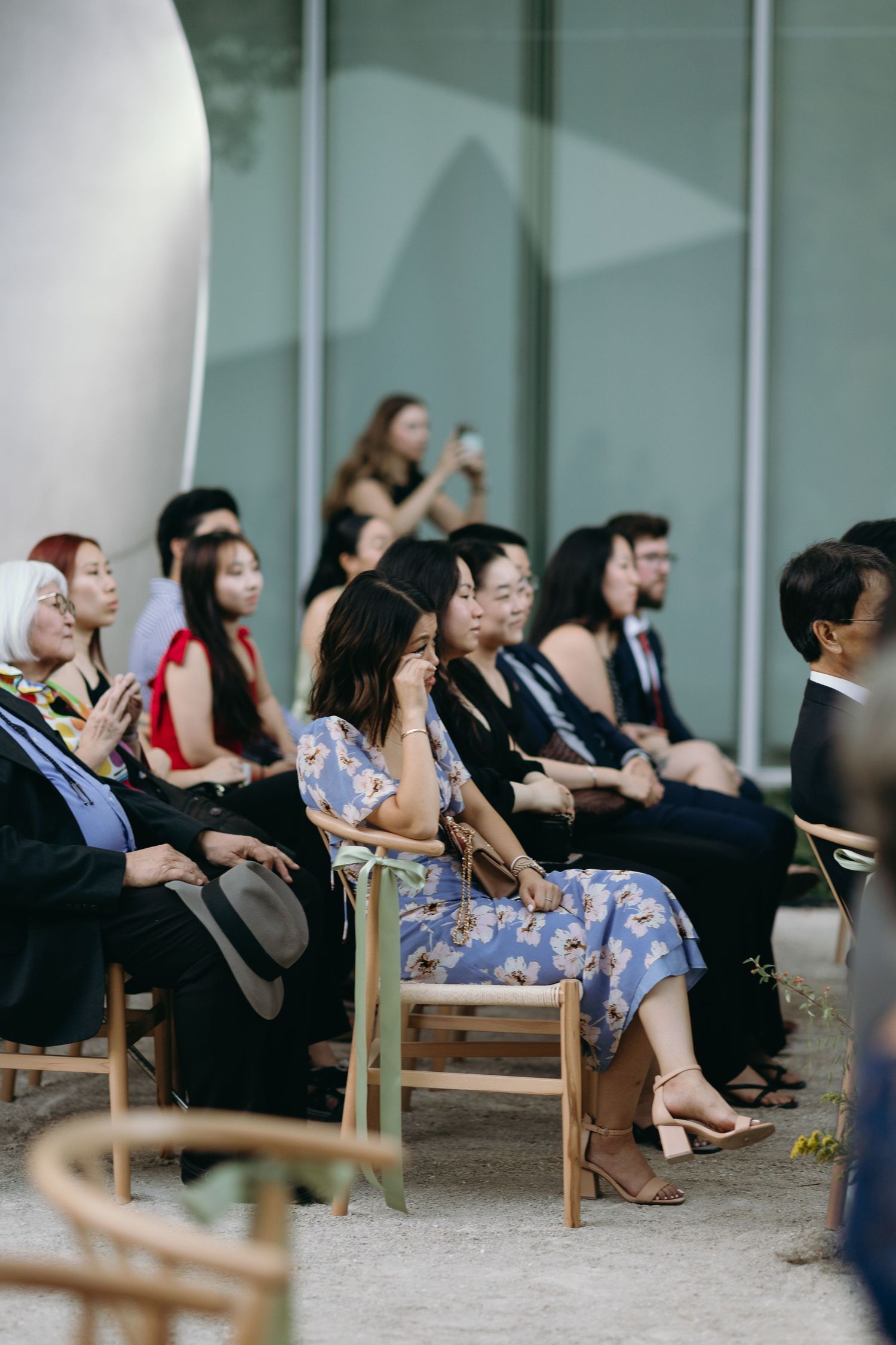 Wedding guests seated on wooden chairs watch the ceremony outdoors.