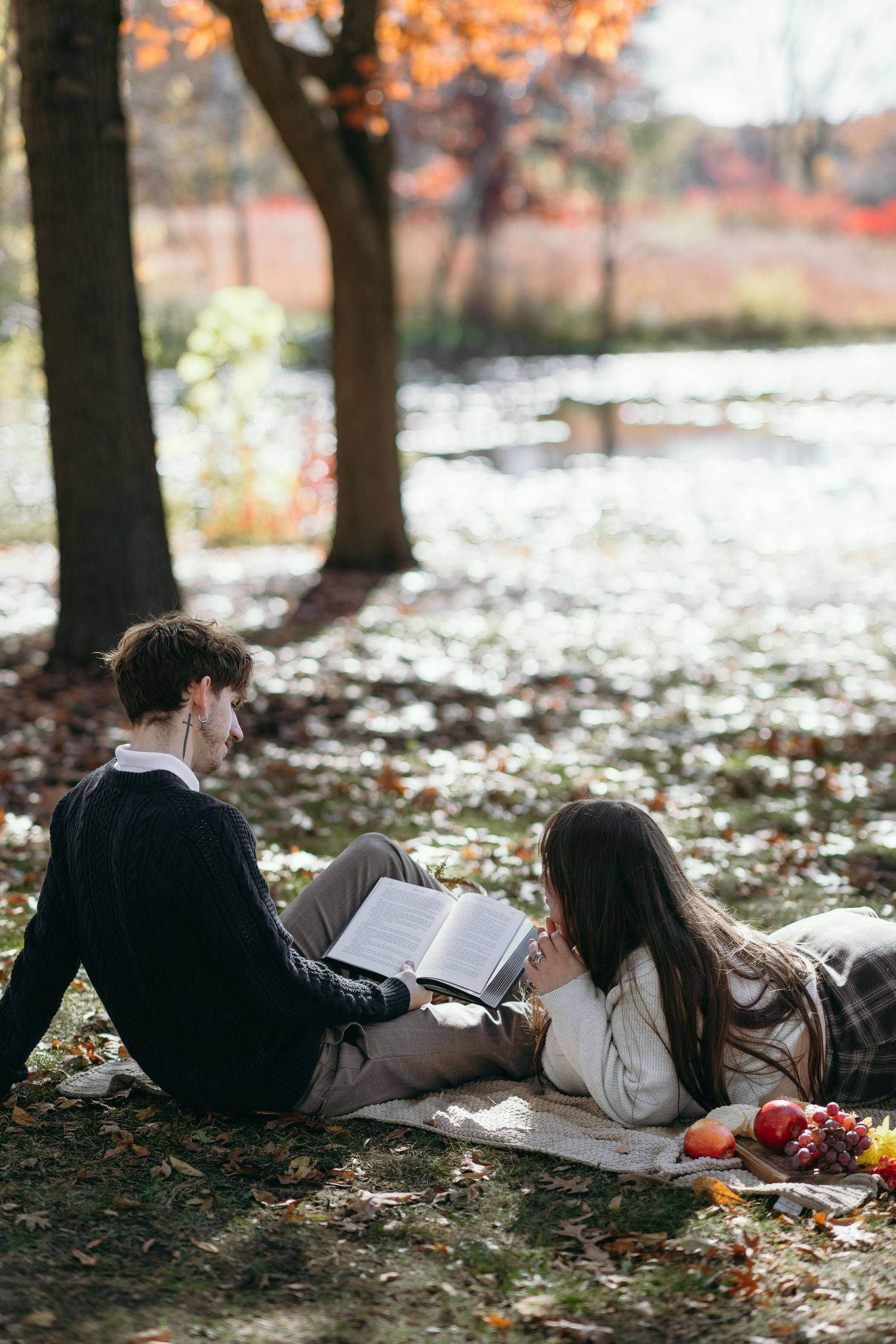 A couple lies on autumn leaves while reading a book together outdoors.