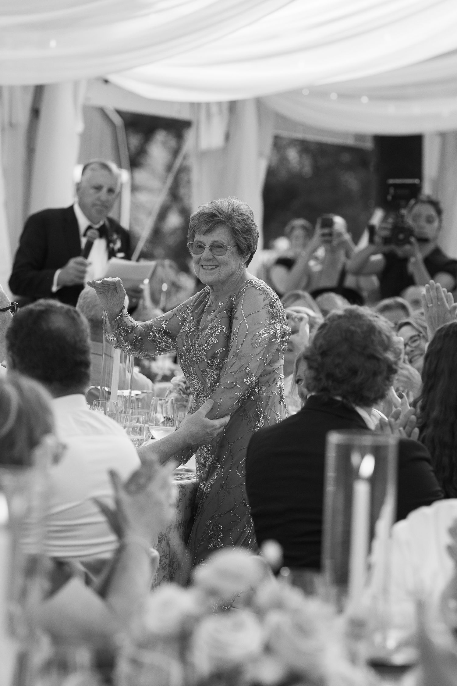 Black and white photo of smiling woman in sequined gown raising glass during wedding toast.