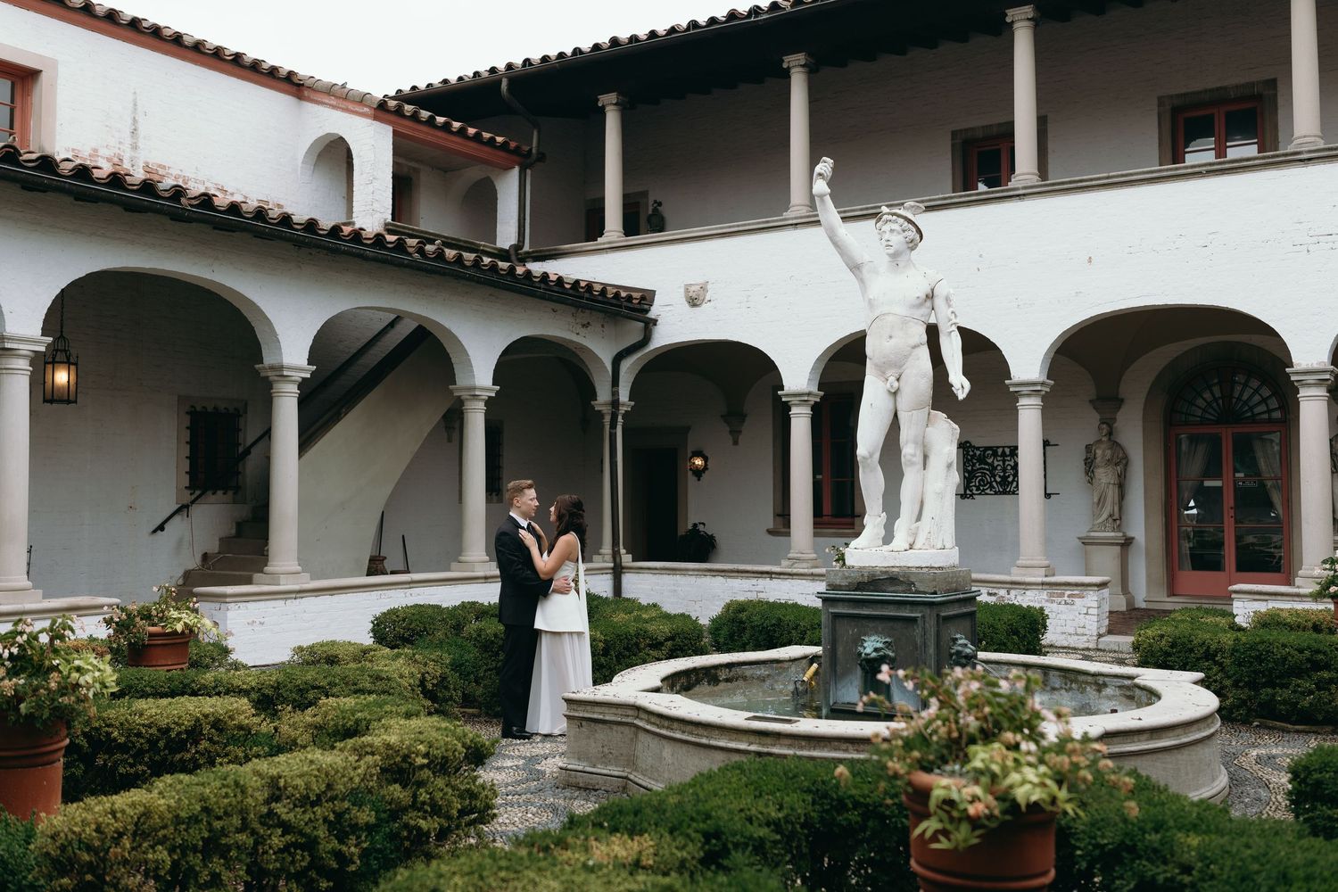 A classic Mediterranean courtyard features a grand white statue centerpiece surrounded by arched columns and manicured gardens.