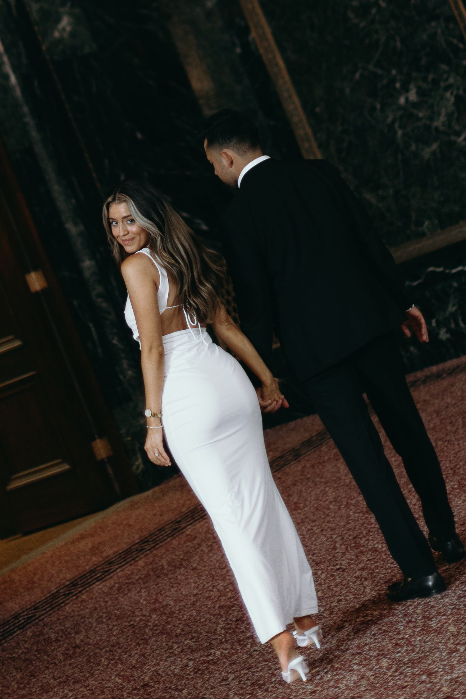 A couple in formal attire at a wedding reception, with the bride wearing a fitted white backless gown.