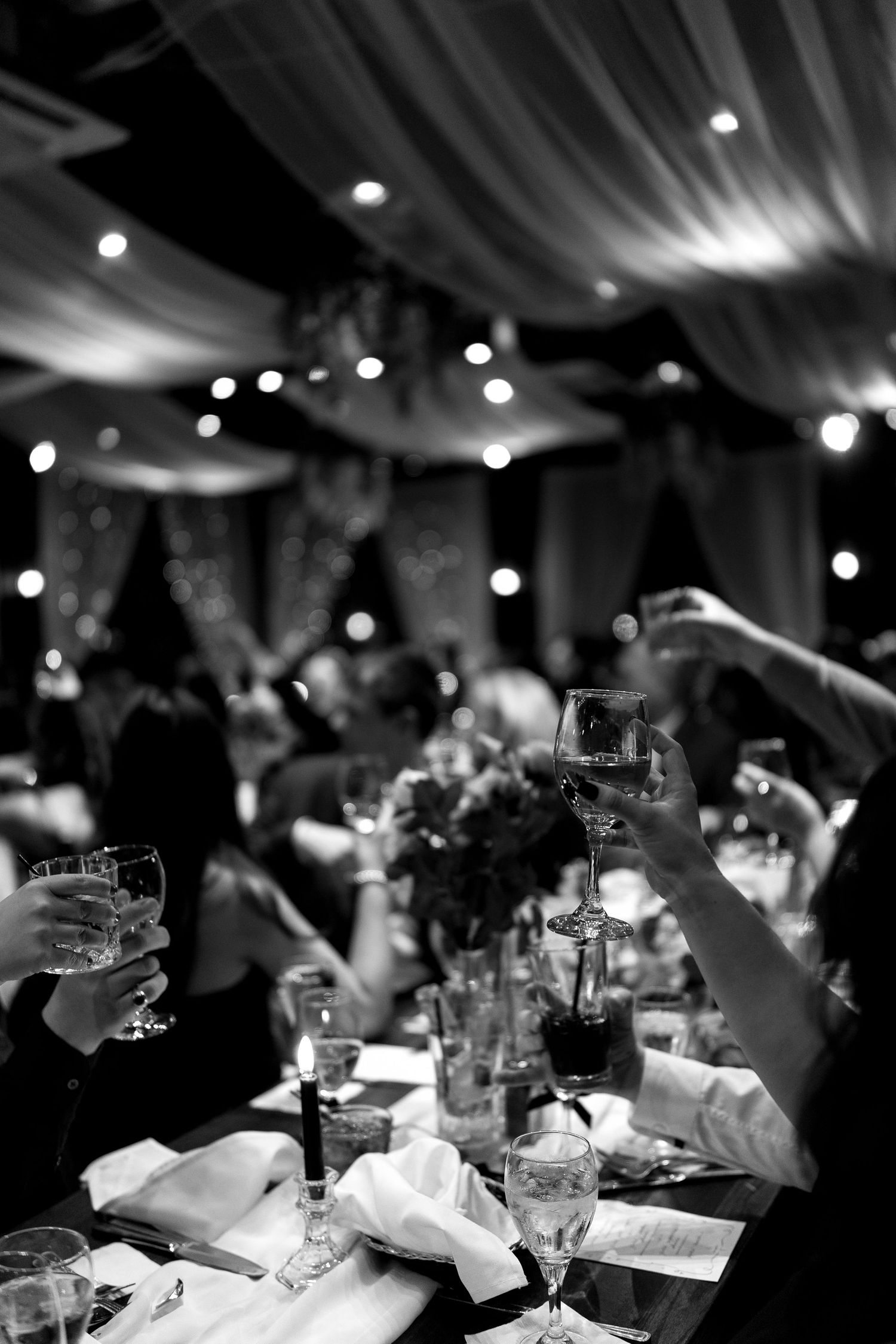 Black and white photo of wedding guests raising glasses for a toast under draped ceiling.