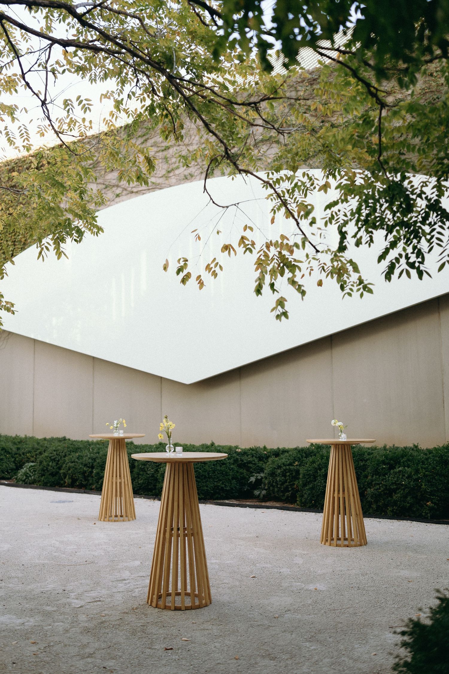 Three modern wooden cocktail tables arranged outdoors under tree branches.