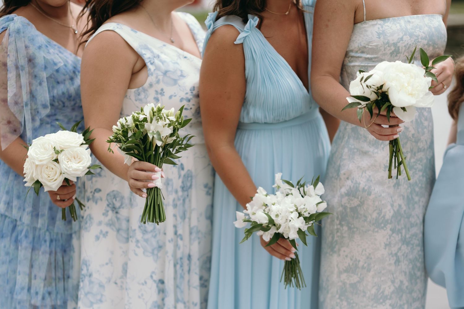 Bridesmaids in light blue dresses stand in line holding white bouquets against a soft floral backdrop.