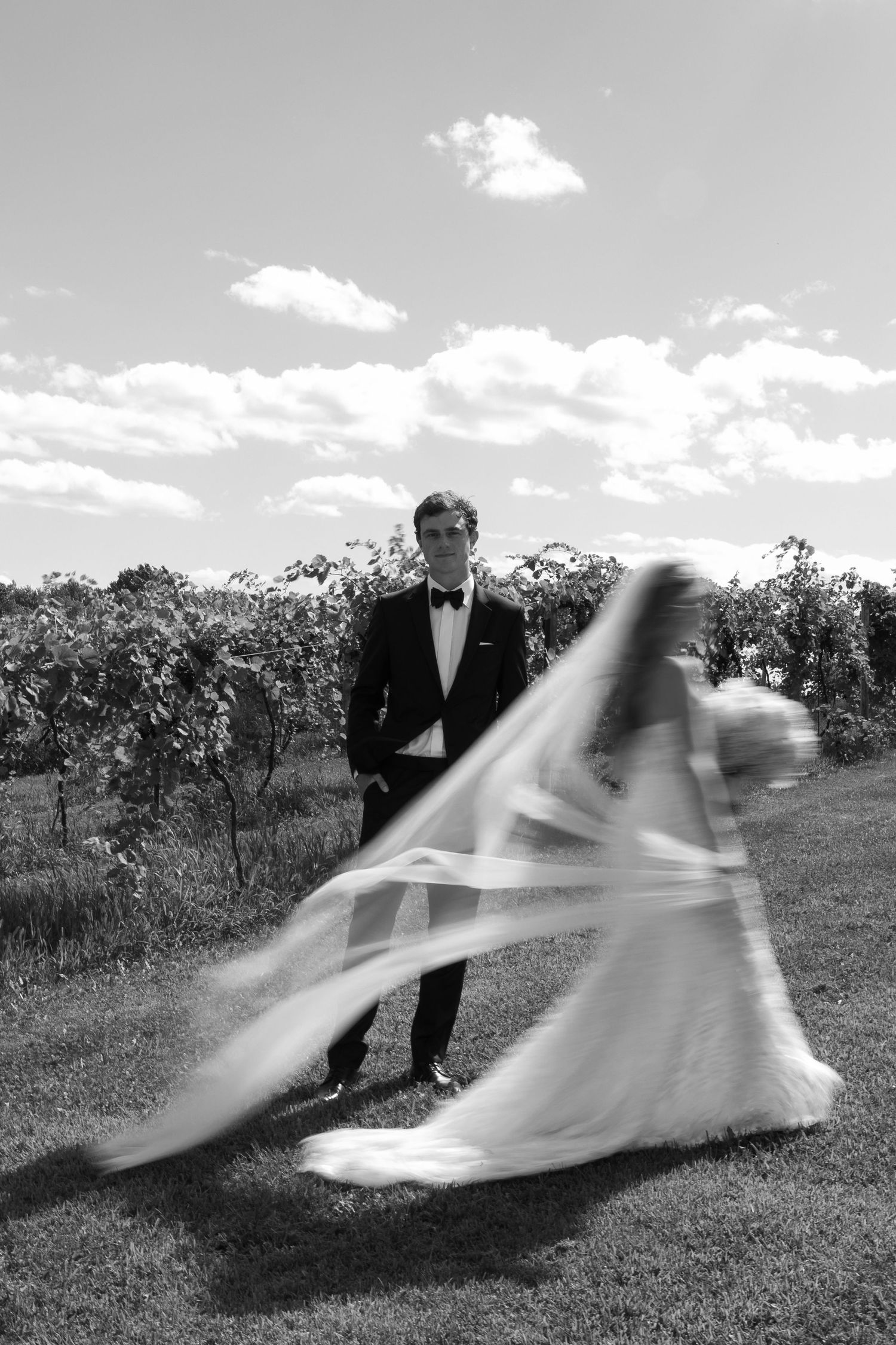 Black and white wedding photo showing a bride in motion with flowing dress and veil next to groom in tuxedo outdoors.