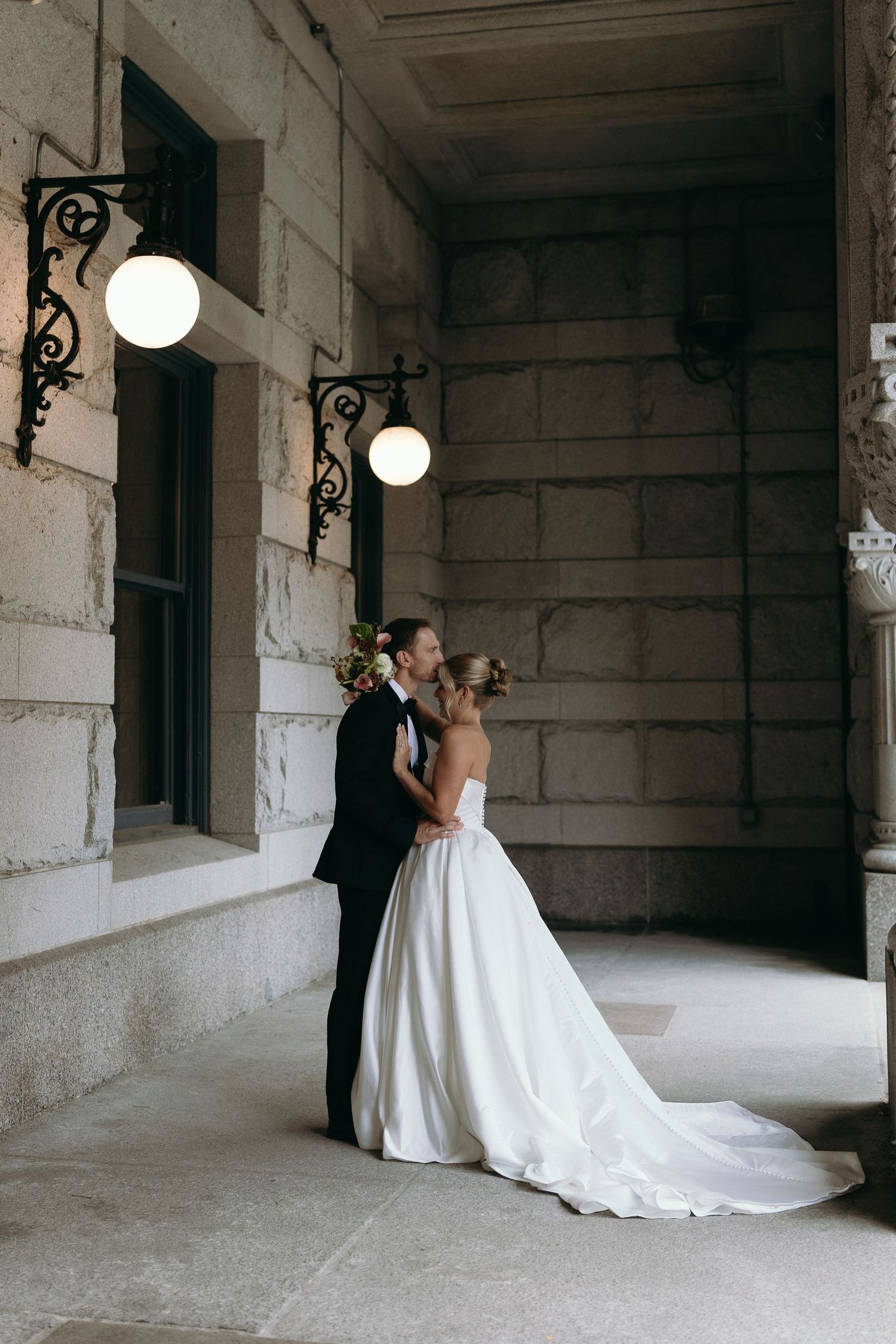 A romantic wedding couple embraces in an elegant stone corridor with white globe lights and arched windows.