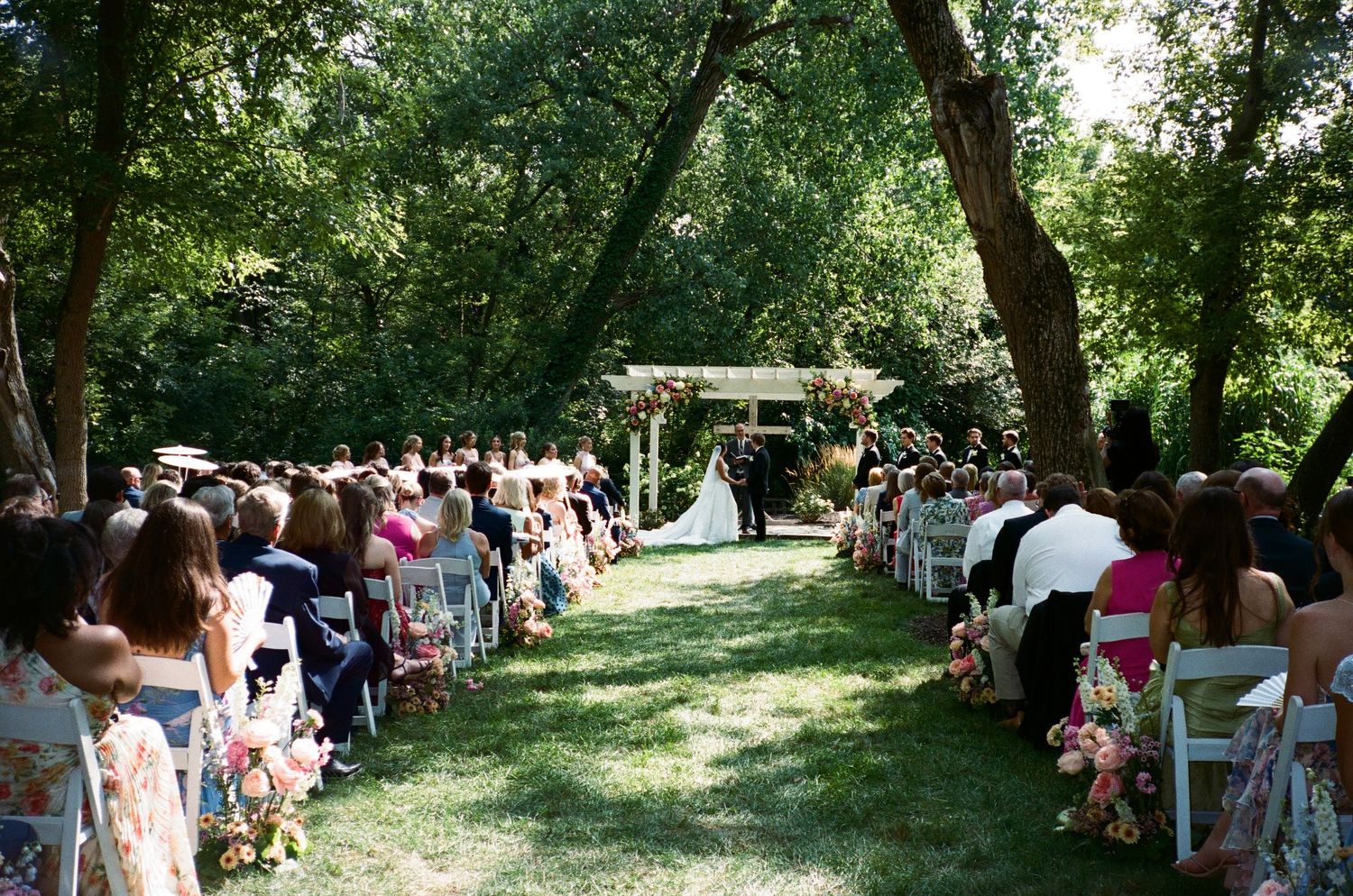 Outdoor wedding ceremony with guests seated on white chairs along a grass aisle leading to a floral arch amid lush trees.