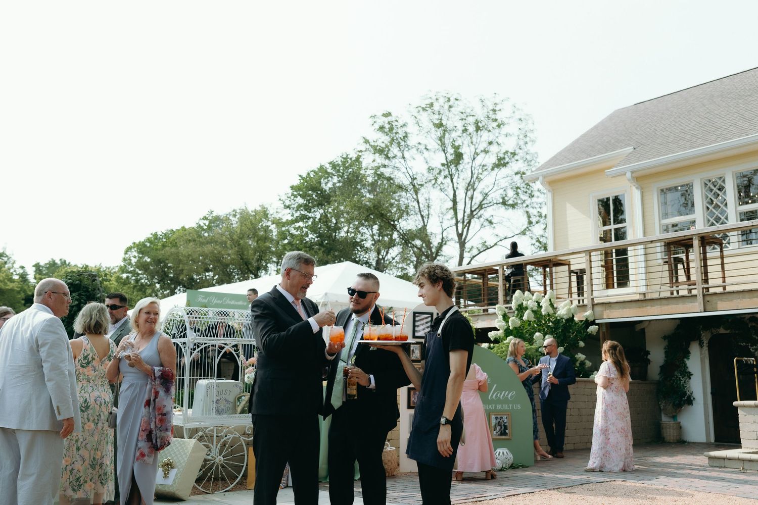 Guests mingle outdoors at an elegant wedding reception near a white carriage and classic house venue.