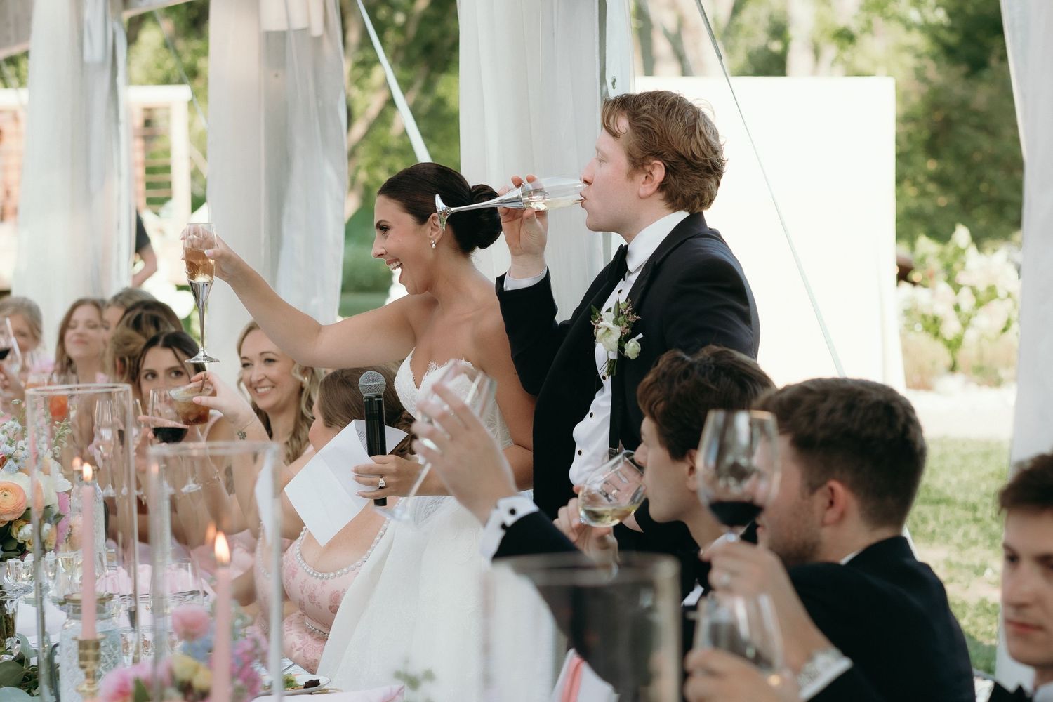 Bride and groom toast at elegant outdoor wedding reception with guests raising glasses under white tent.