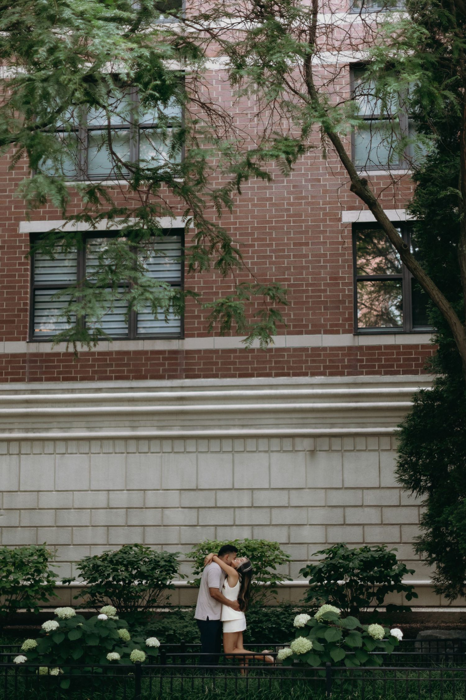 A couple shares a tender moment in front of a brick building with white accents and flowering bushes.