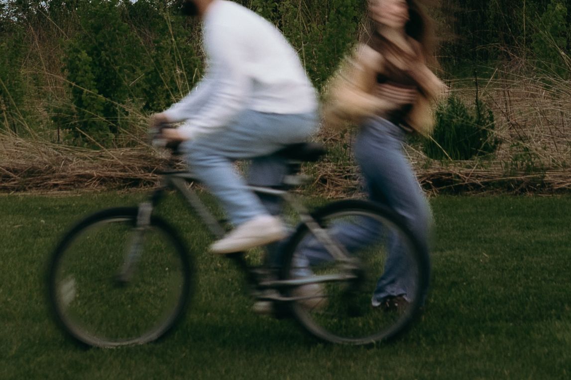 Two people riding a bicycle together on green grass, captured in motion blur near lush trees. Fun unqiue chicago wedding photographer