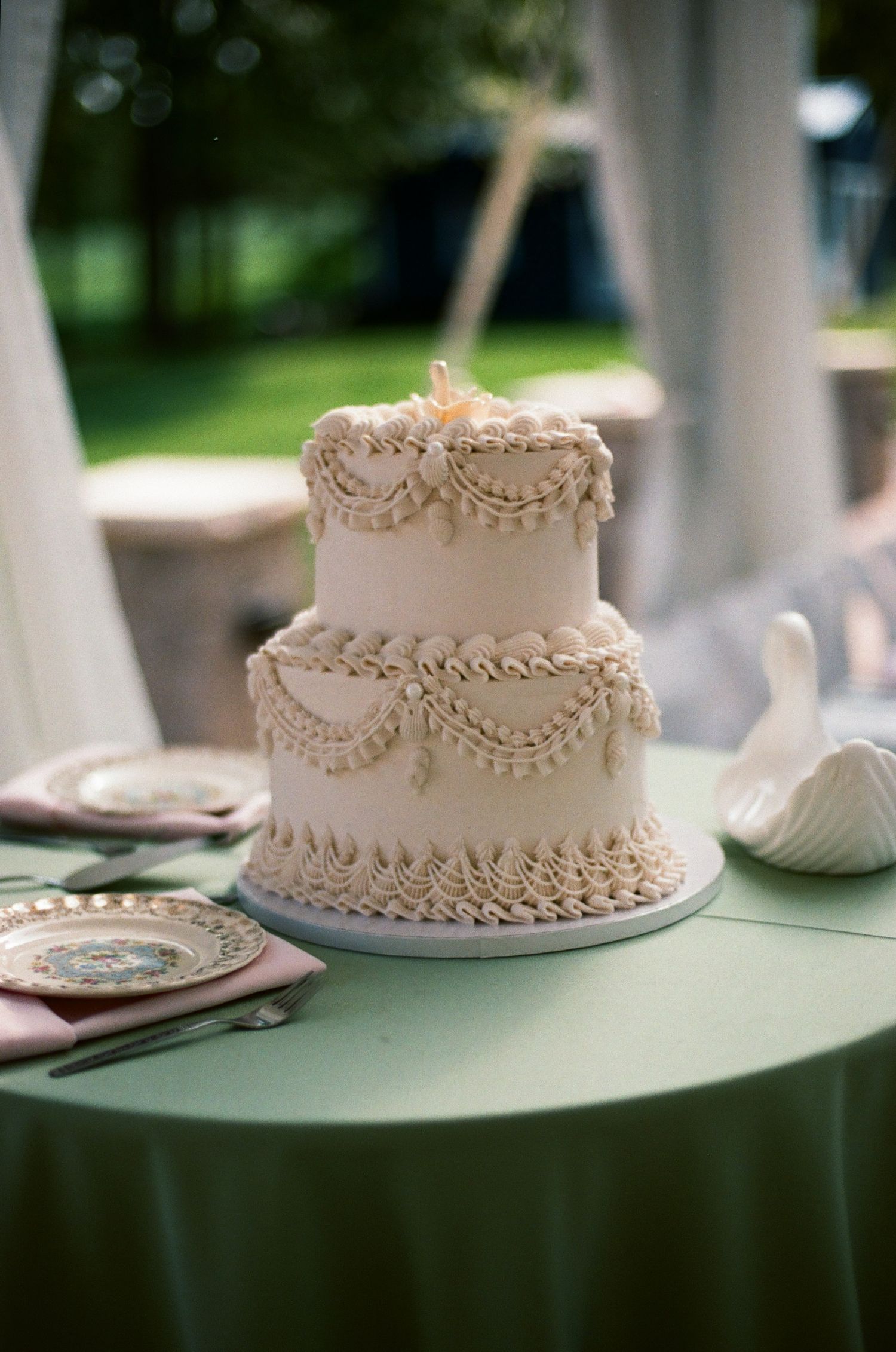 Elegant two-tier white wedding cake with ruffled frosting, swag details, and pearl accents on a green tablecloth.