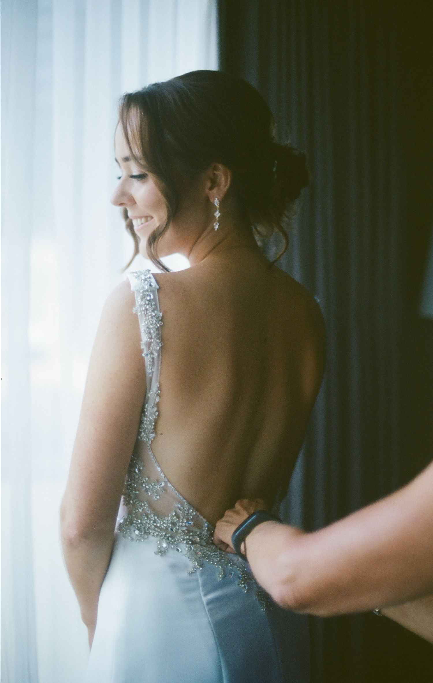 Bride in backless beaded wedding gown smiling as someone fastens her dress near a window.