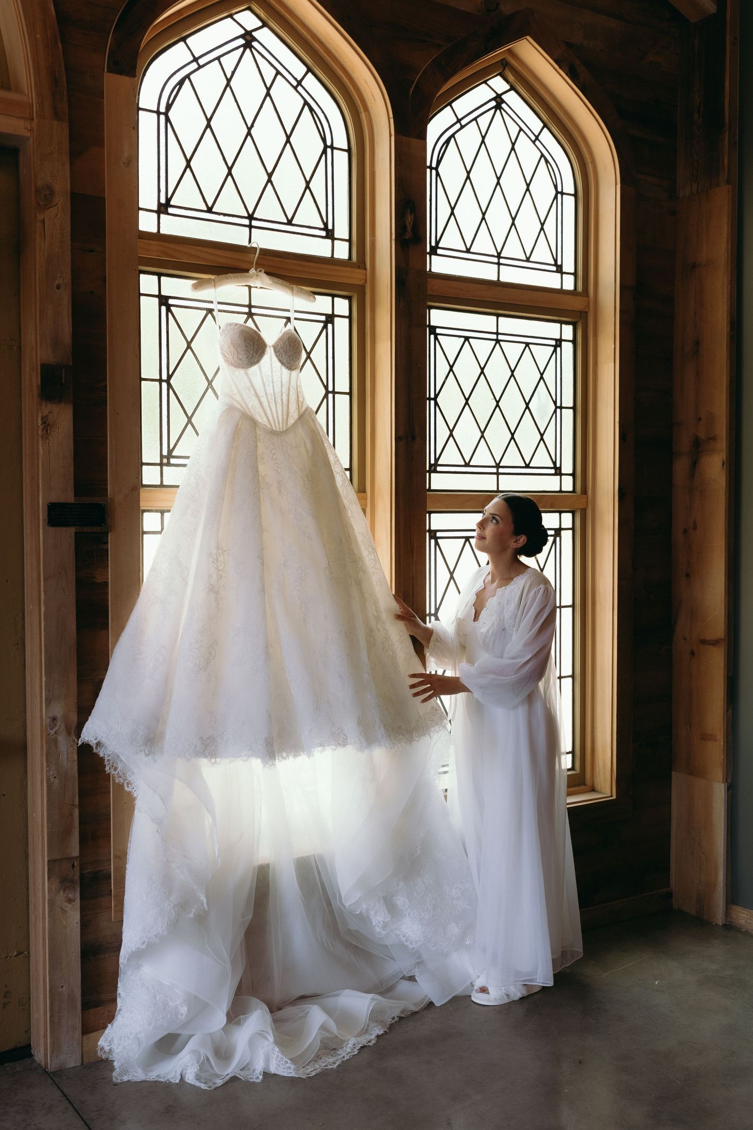 Bride in white robe admiring her wedding gown hanging by arched stained glass windows in elegant venue.
