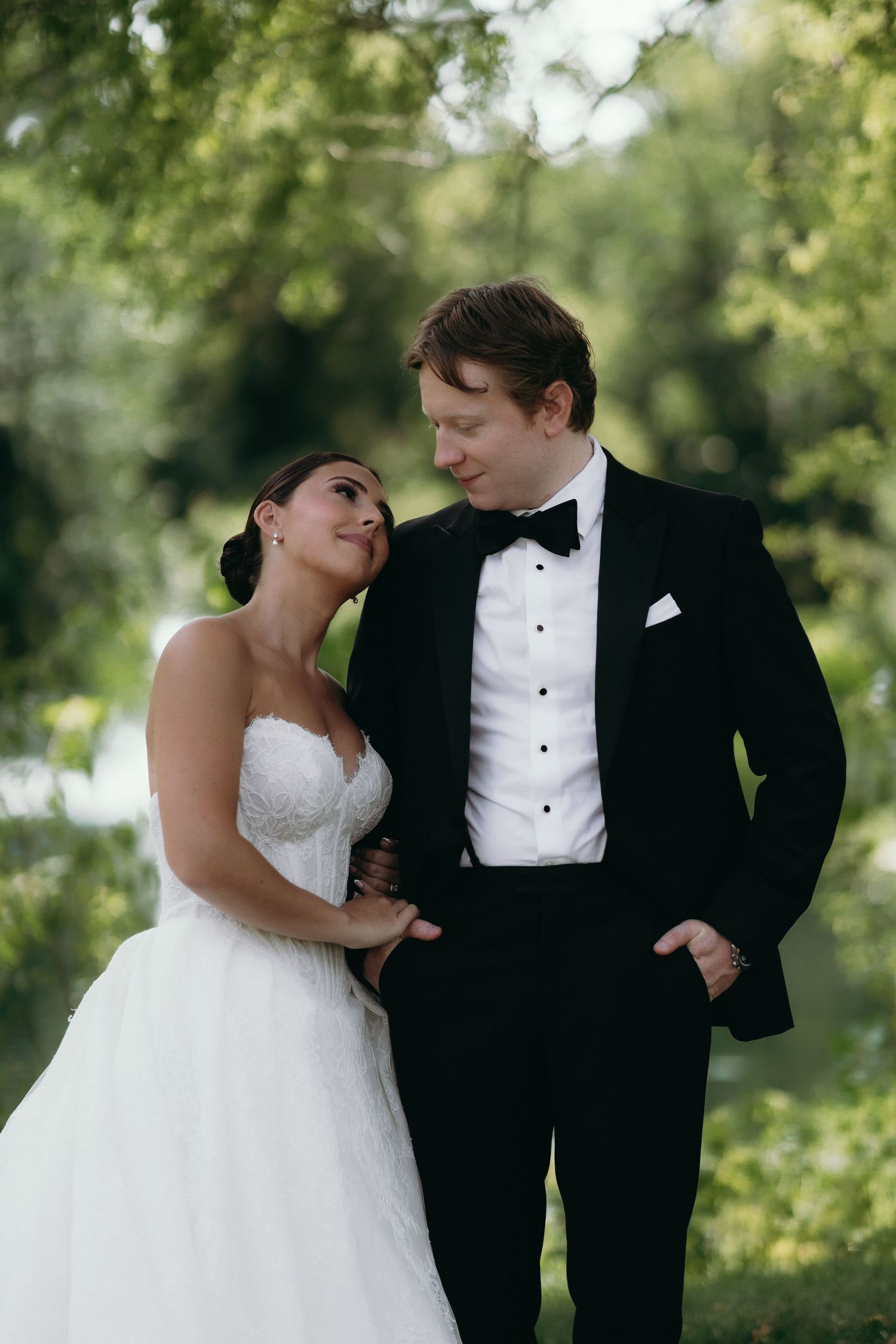 Bride in white gown and groom in black tuxedo share a tender moment outdoors surrounded by lush green trees.