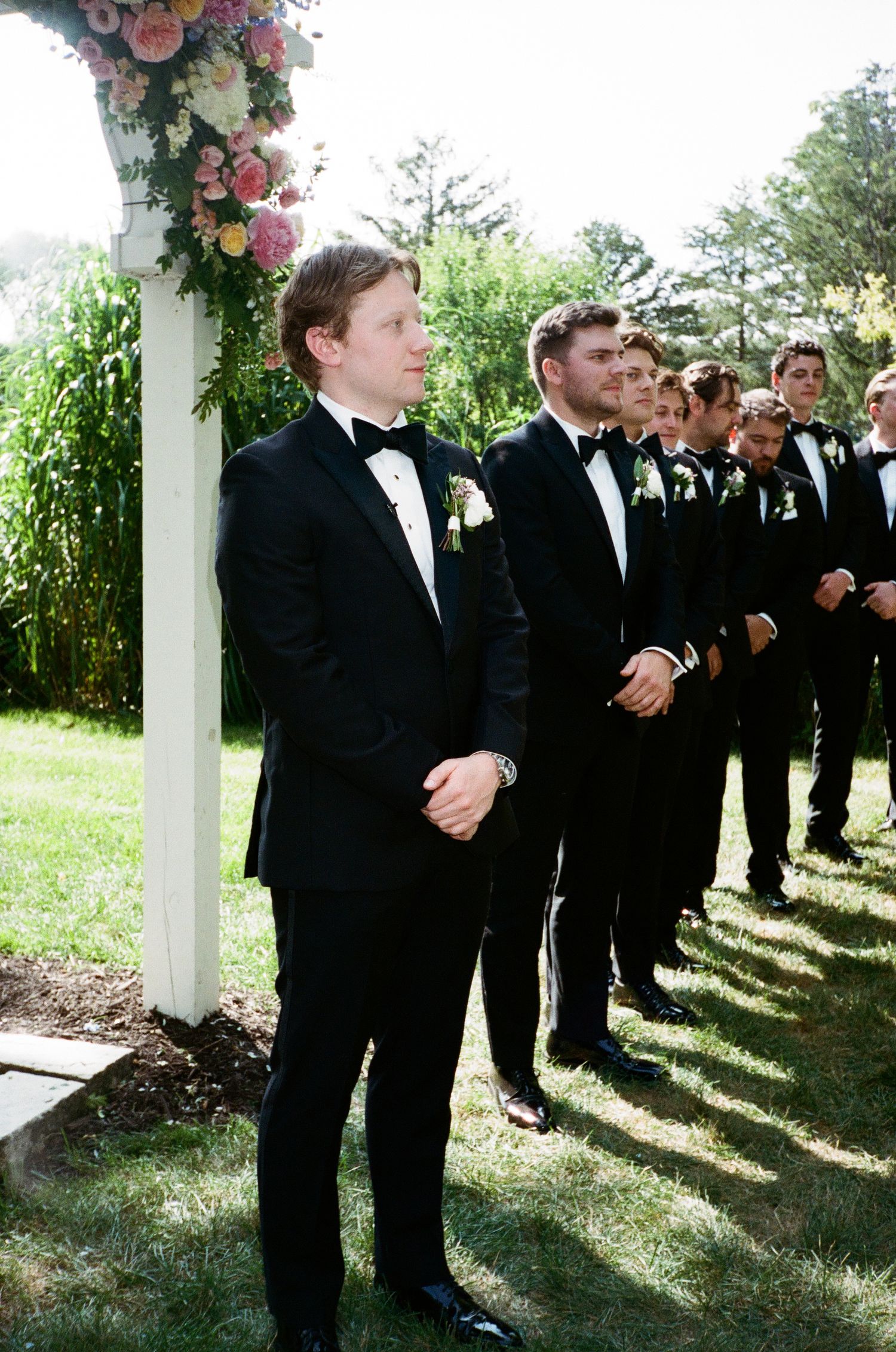 Groomsmen in black tuxedos with boutonnieres stand at outdoor wedding ceremony beside floral arch.