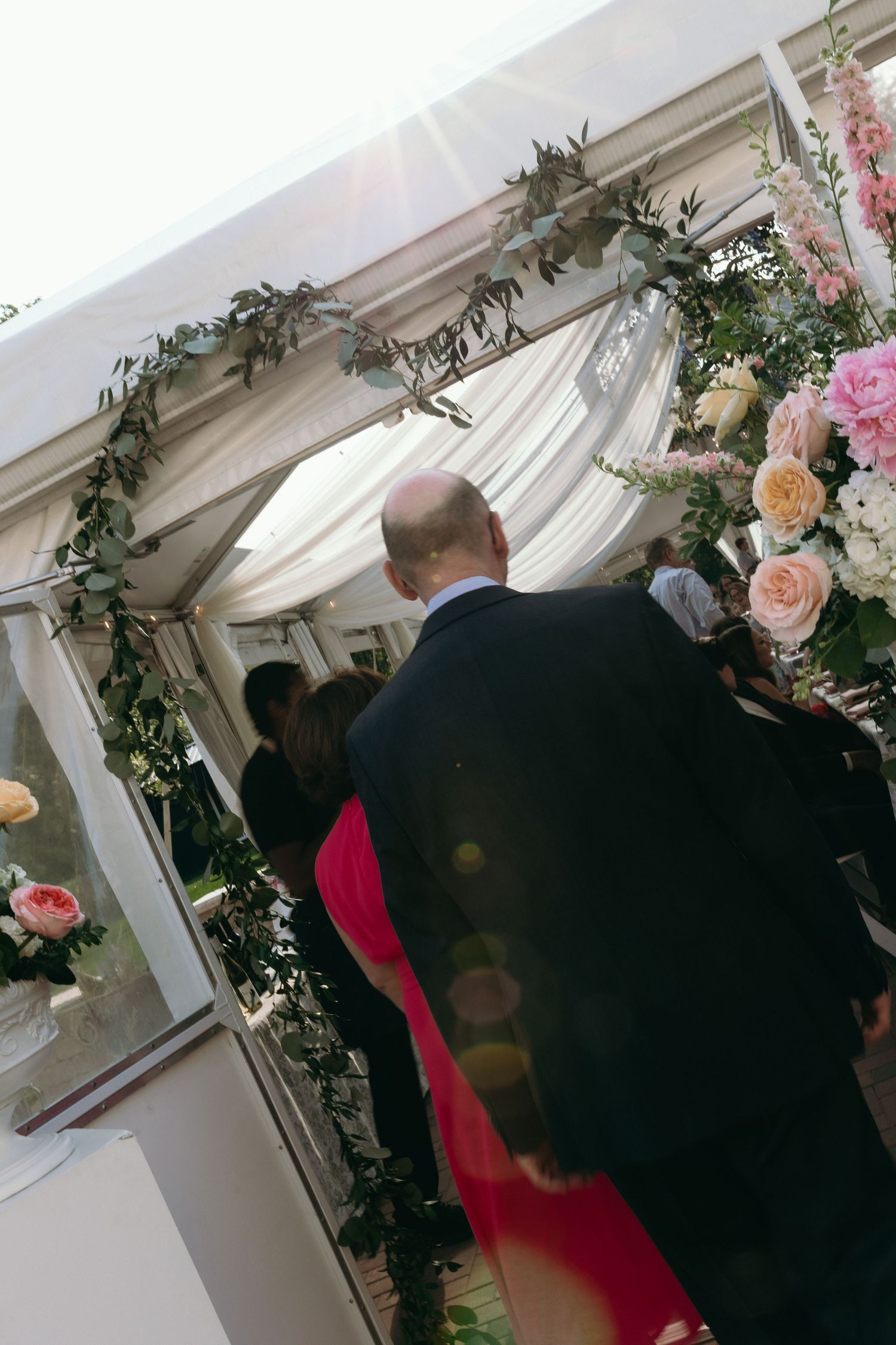 Bald man in black suit enters floral arch wedding tent with pink roses and greenery garland.