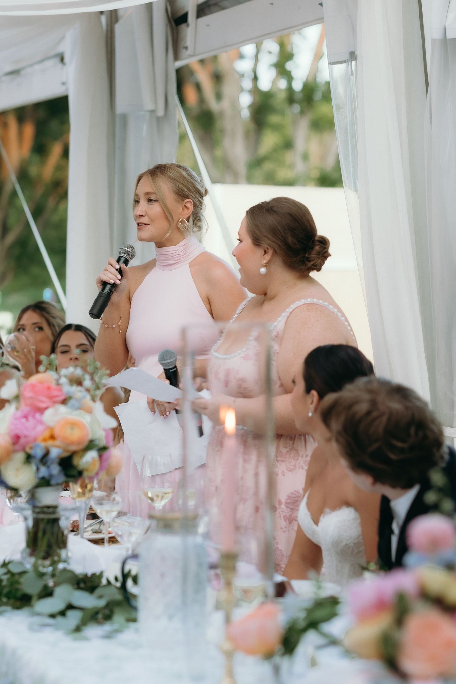 Bridesmaid in pink dress giving wedding speech with microphone at floral head table.