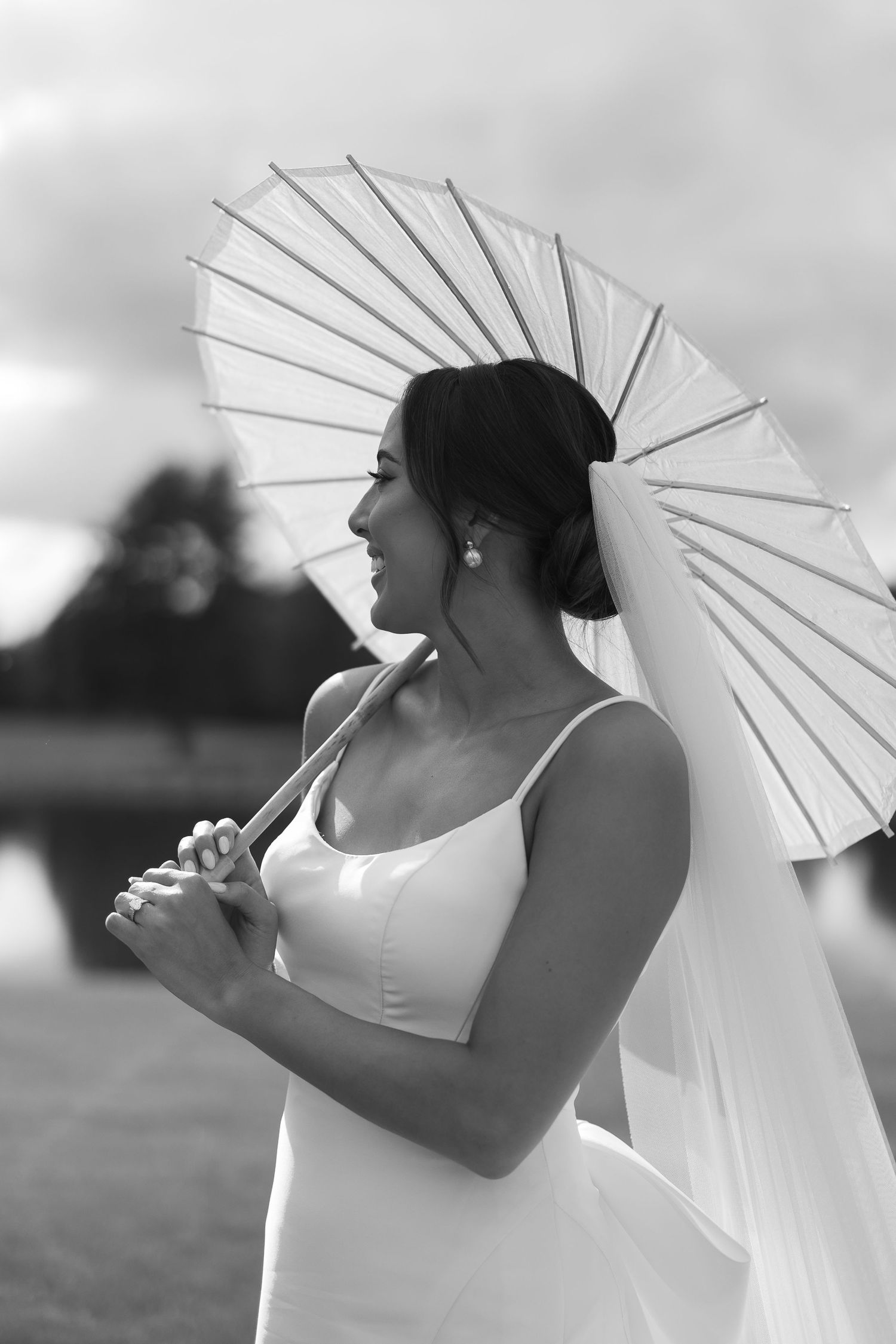 A bride in a sleek white dress holds a delicate white parasol in a black and white portrait.