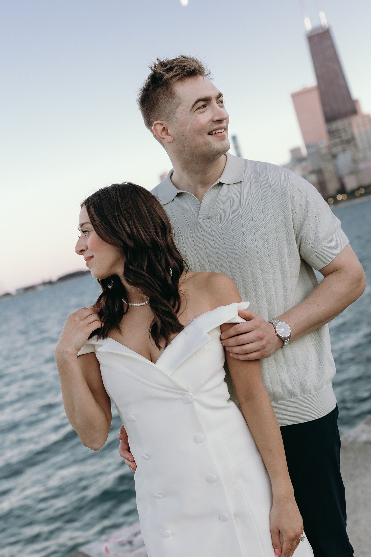 A couple embraces by Lake Michigan at sunset with the Chicago skyline and Willis Tower visible in the background.
