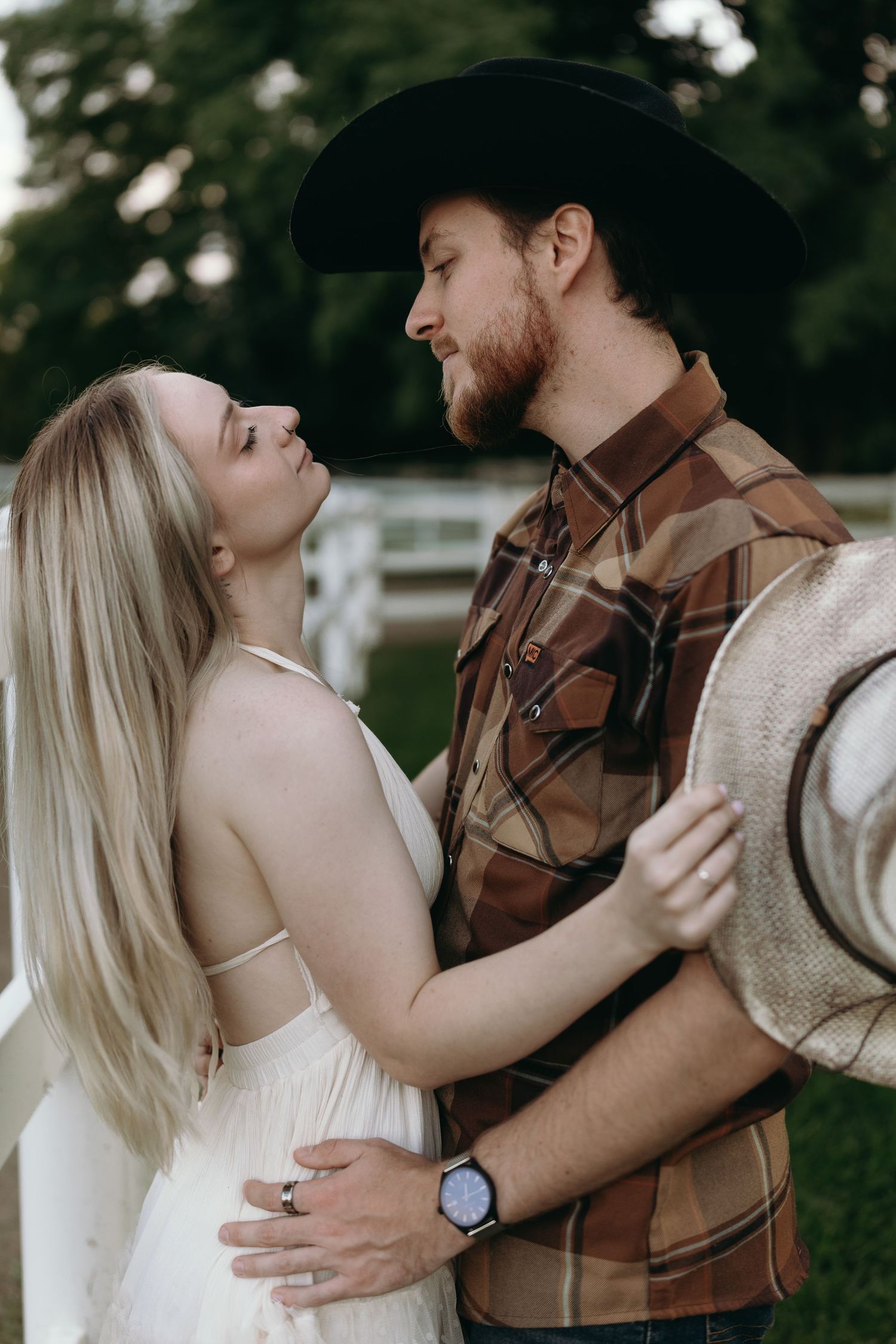 A romantic couple embraces outdoors while the person on the right wears a plaid shirt and cowboy hat in a rustic setting.