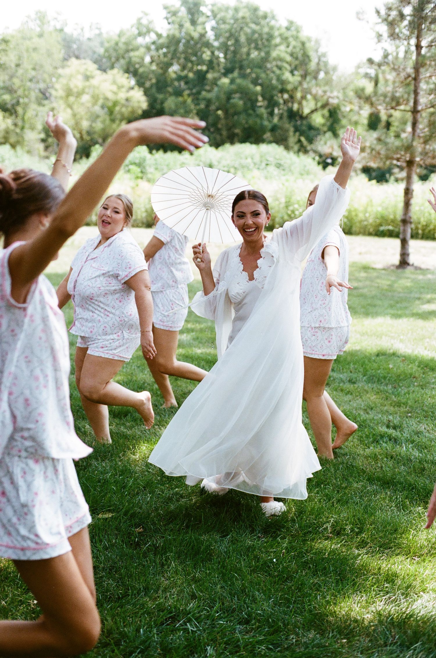 A joyful bride in a white gown twirls with parasol among bridesmaids in matching white pajamas on a lush green lawn.
