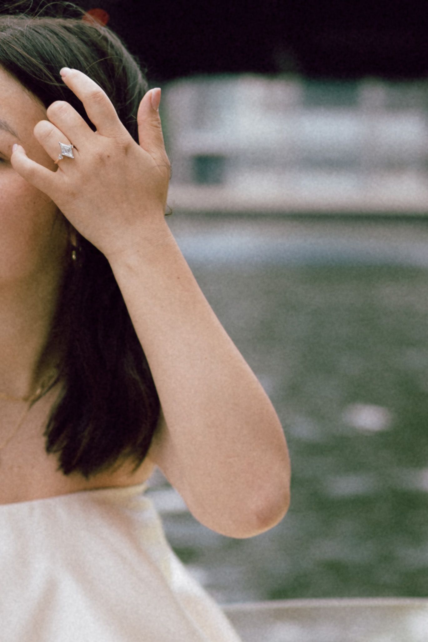 Woman in white dress wearing a diamond engagement ring, hand raised to dark hair near waterfront.