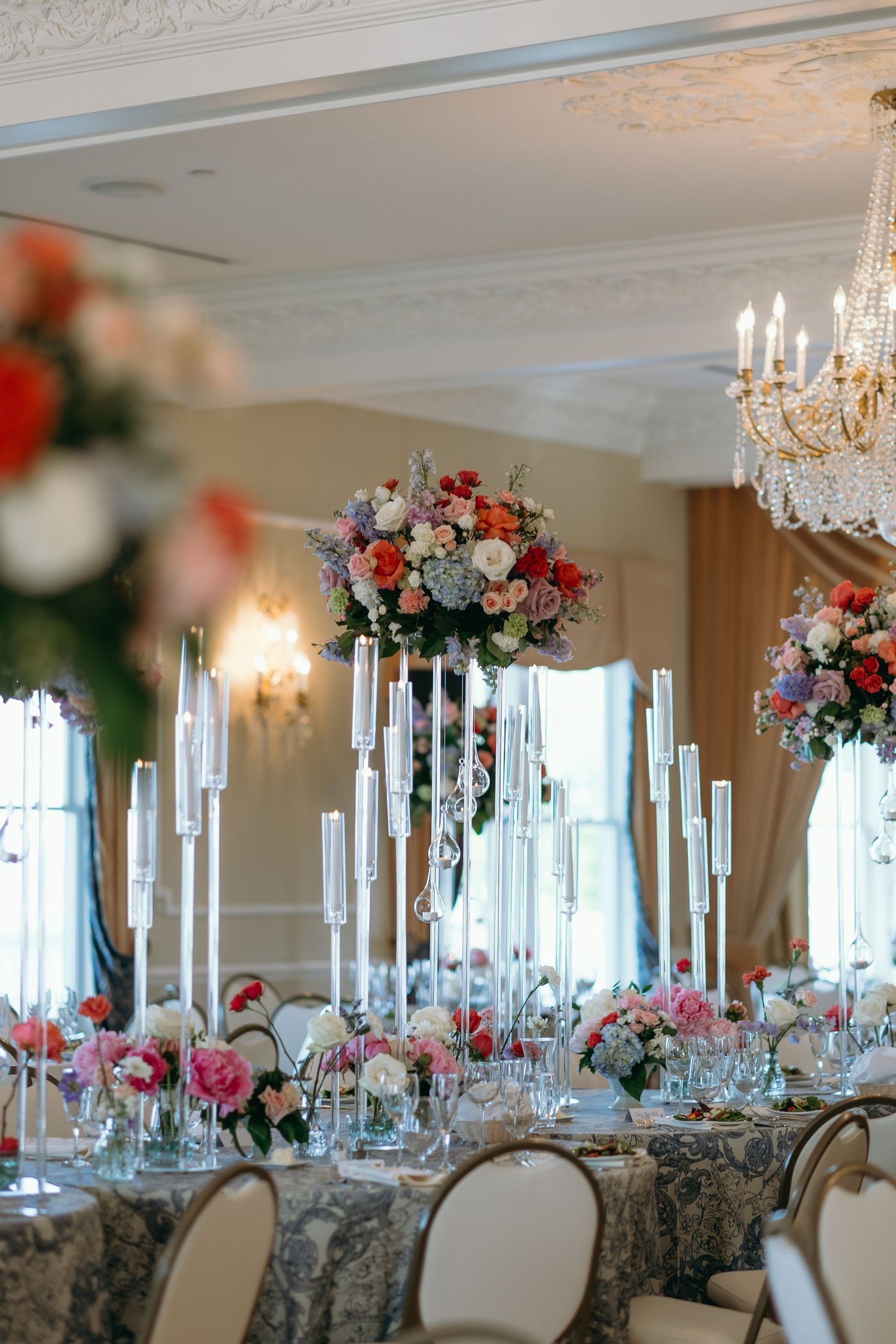 Elegant wedding reception table with tall floral centerpieces, crystal chandelier, and gray damask linens in luxurious ballroom.