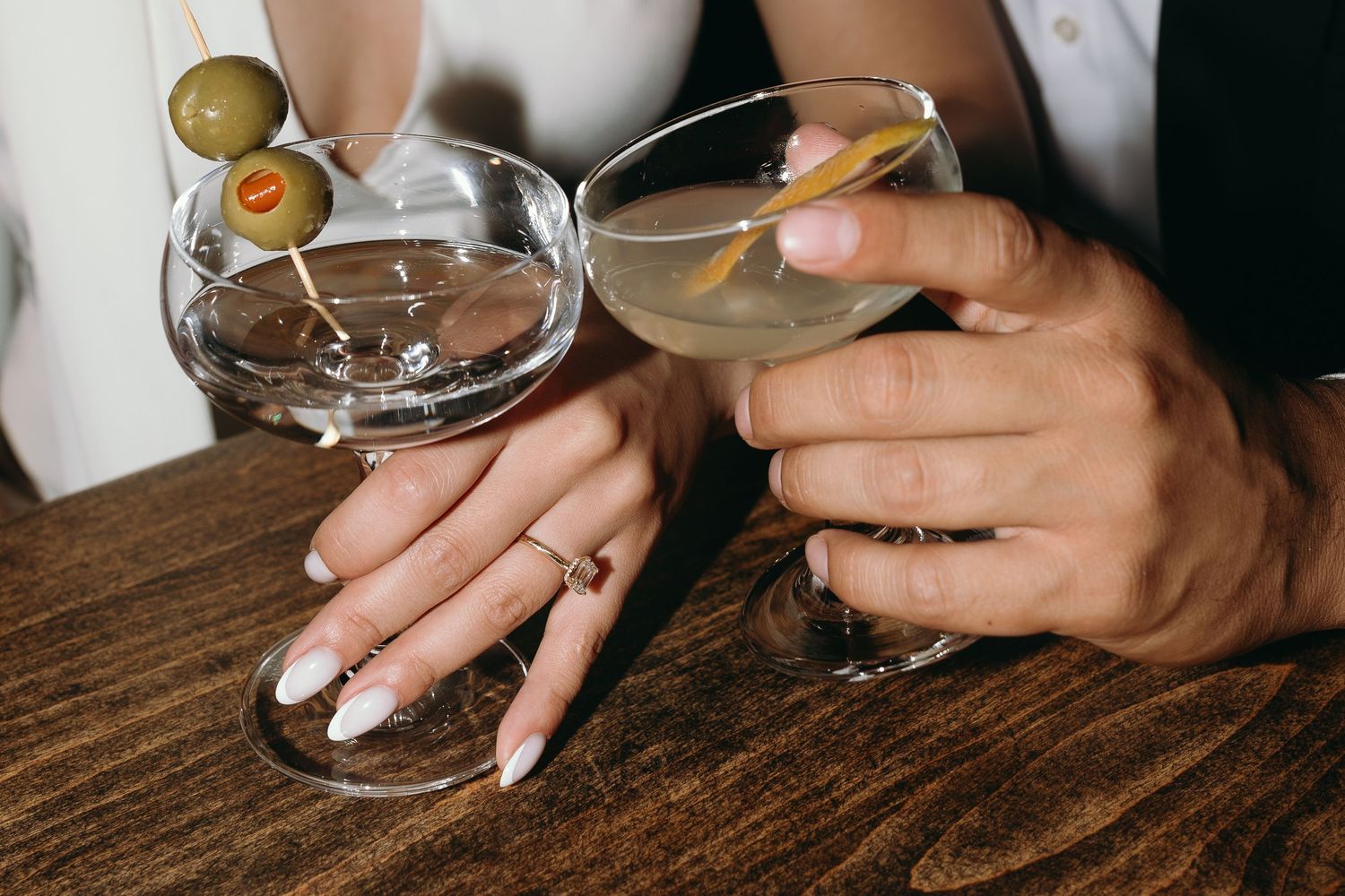 Close up of hands holding martini glasses with olives on wooden table.