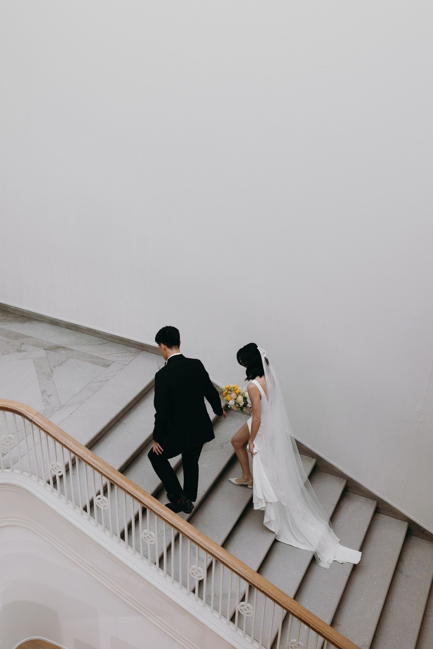 A wedding couple ascending a grand staircase with white railings in formal attire.