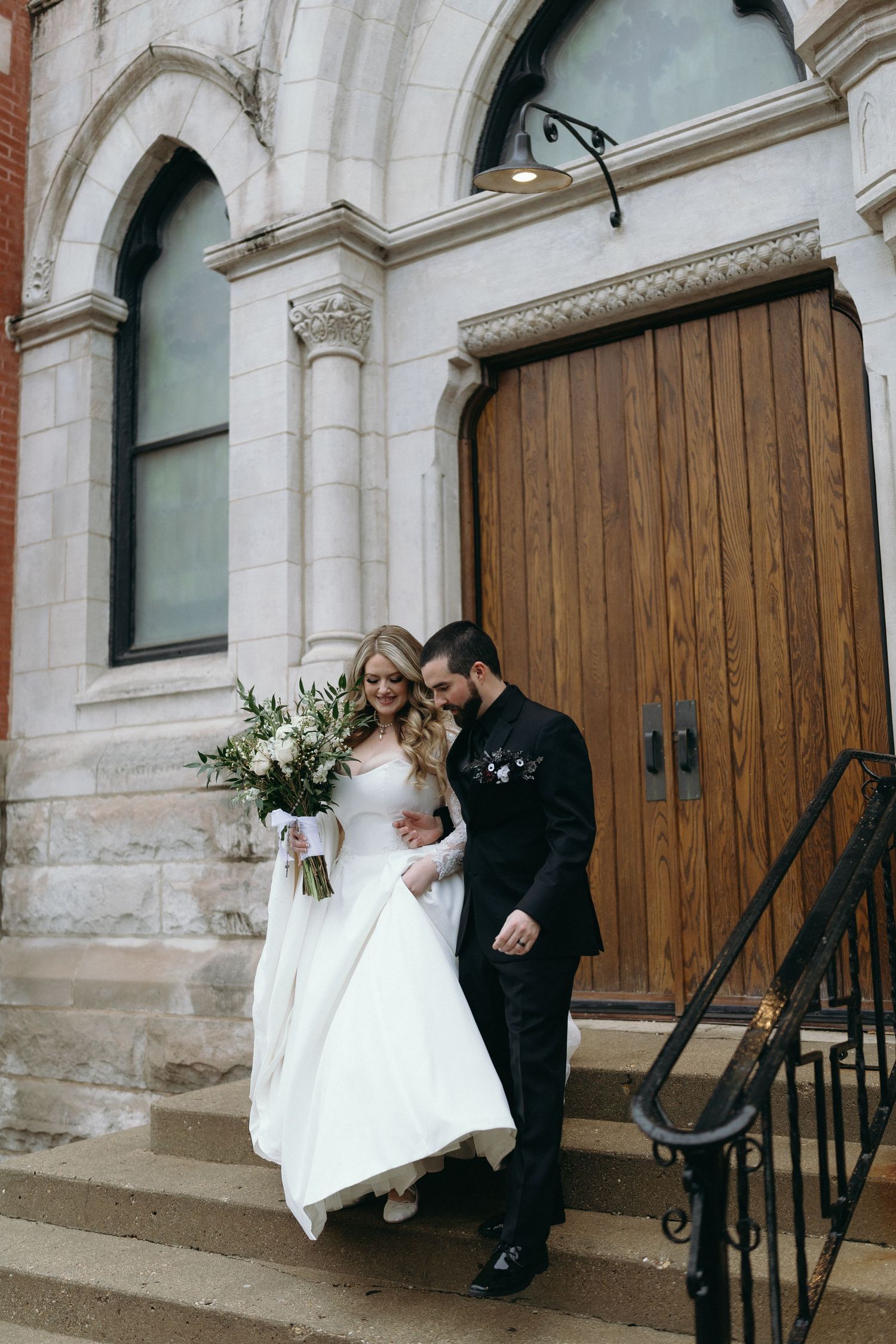 A couple in wedding attire stand together on church steps beside wooden doors.