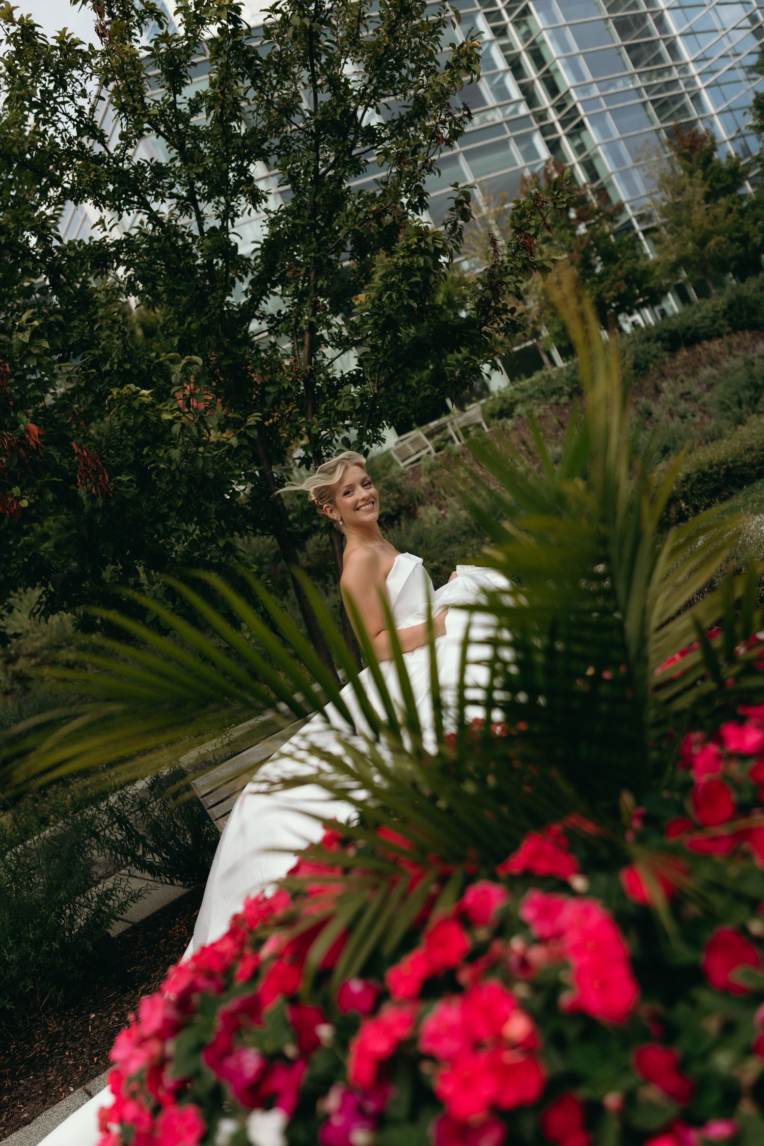 A couple in wedding attire poses in a lush garden setting with vibrant pink flowers and palm fronds in the foreground.
