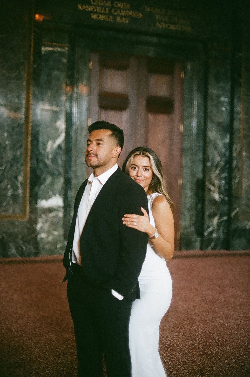 A romantic wedding couple poses together against a dark marble wall in dimly lit elegant interior setting.