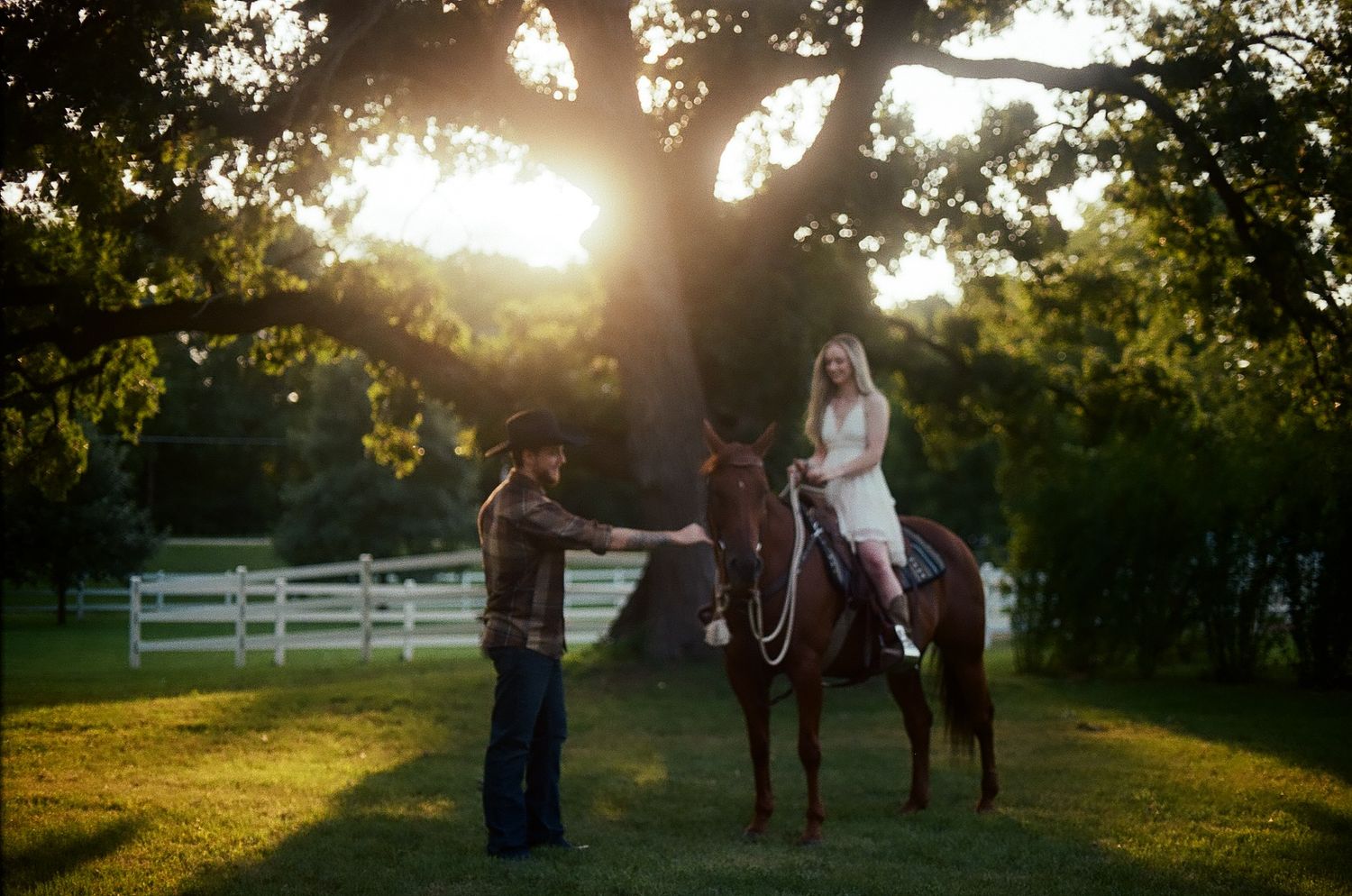Sunset silhouettes a person on horseback and another standing near white fences on a rural farm property.