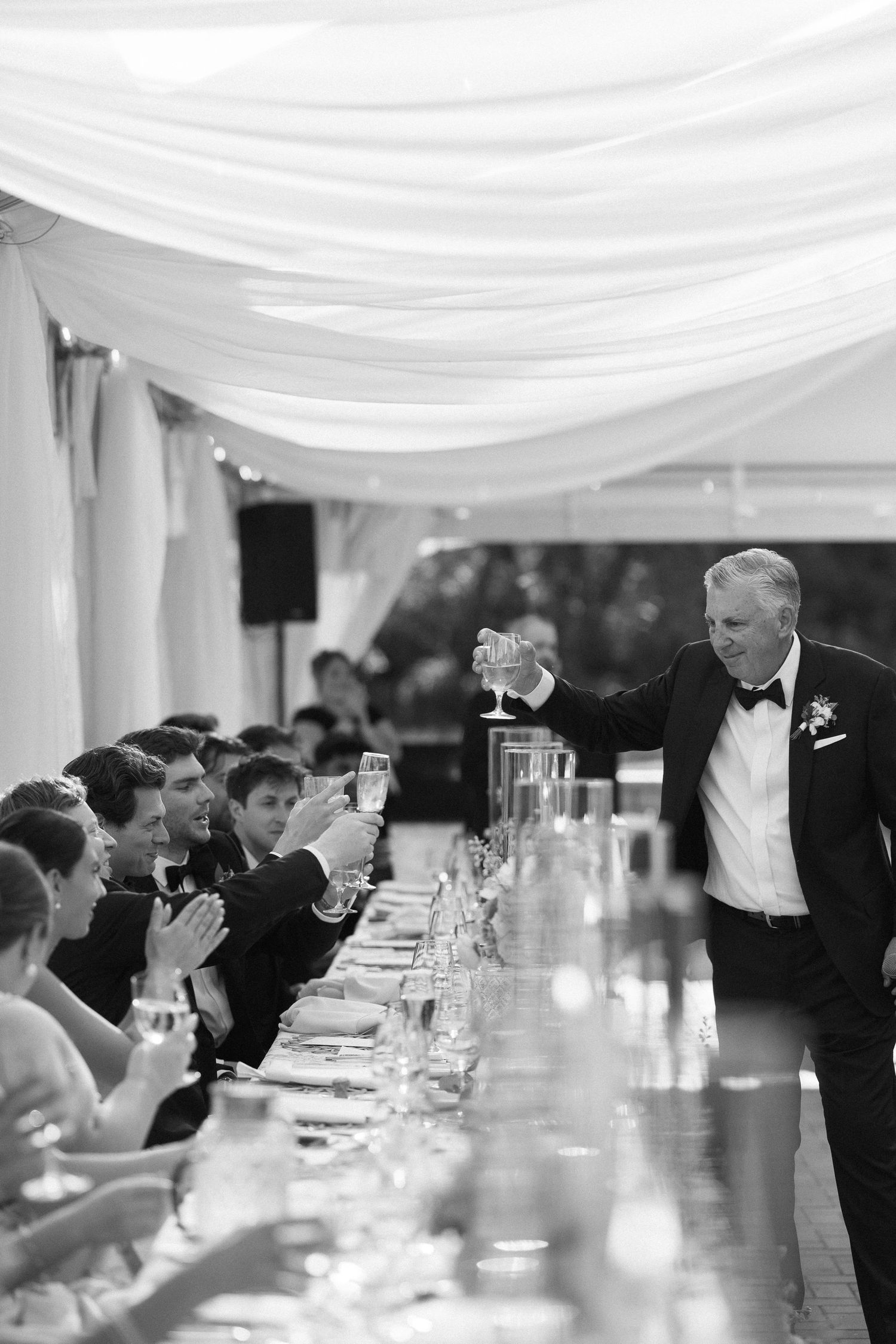 Black and white photo of man in tuxedo toasting wedding party at long reception table.