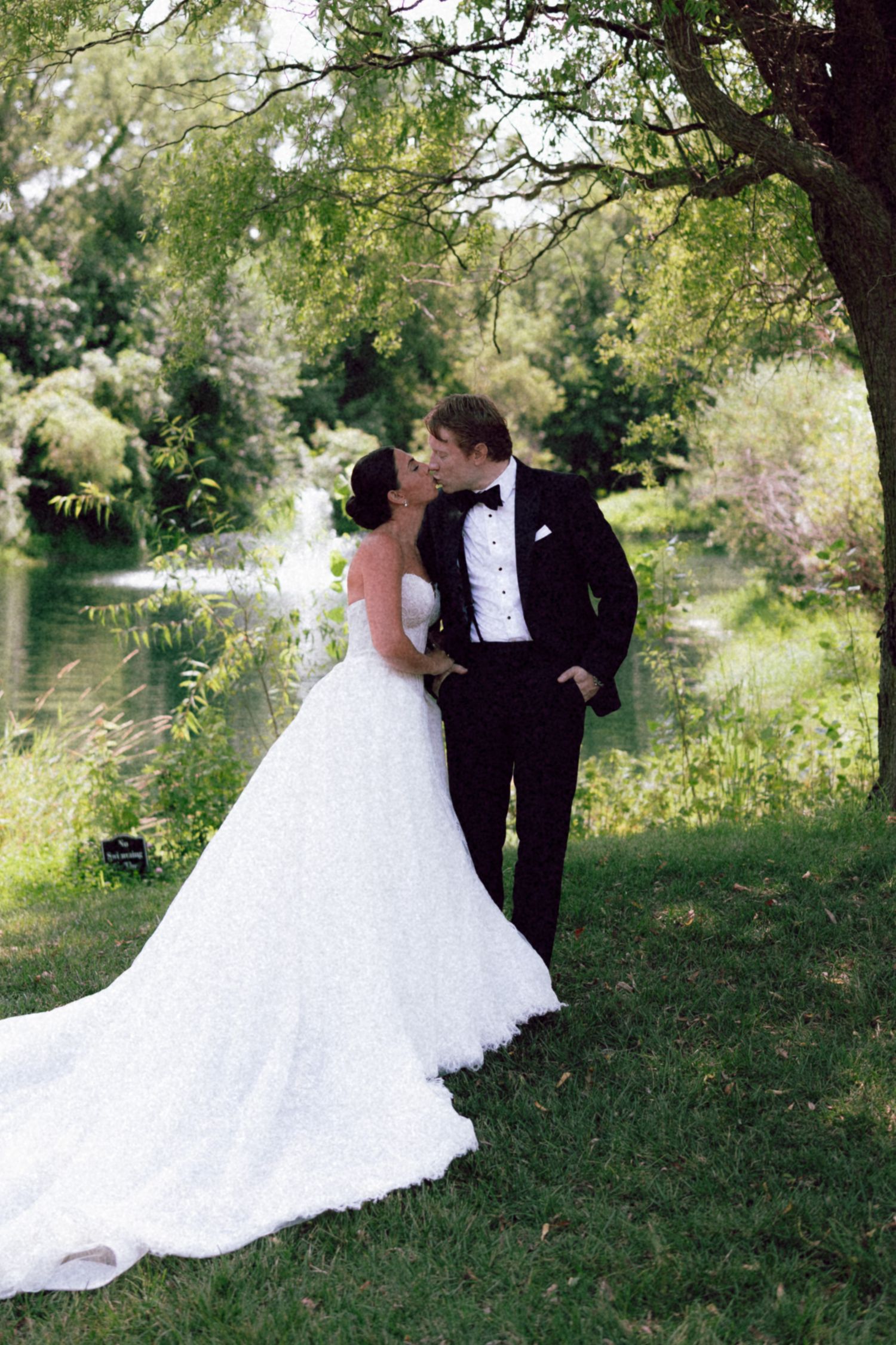 A bride and groom share a romantic kiss under a willow tree beside a serene pond on their wedding day.