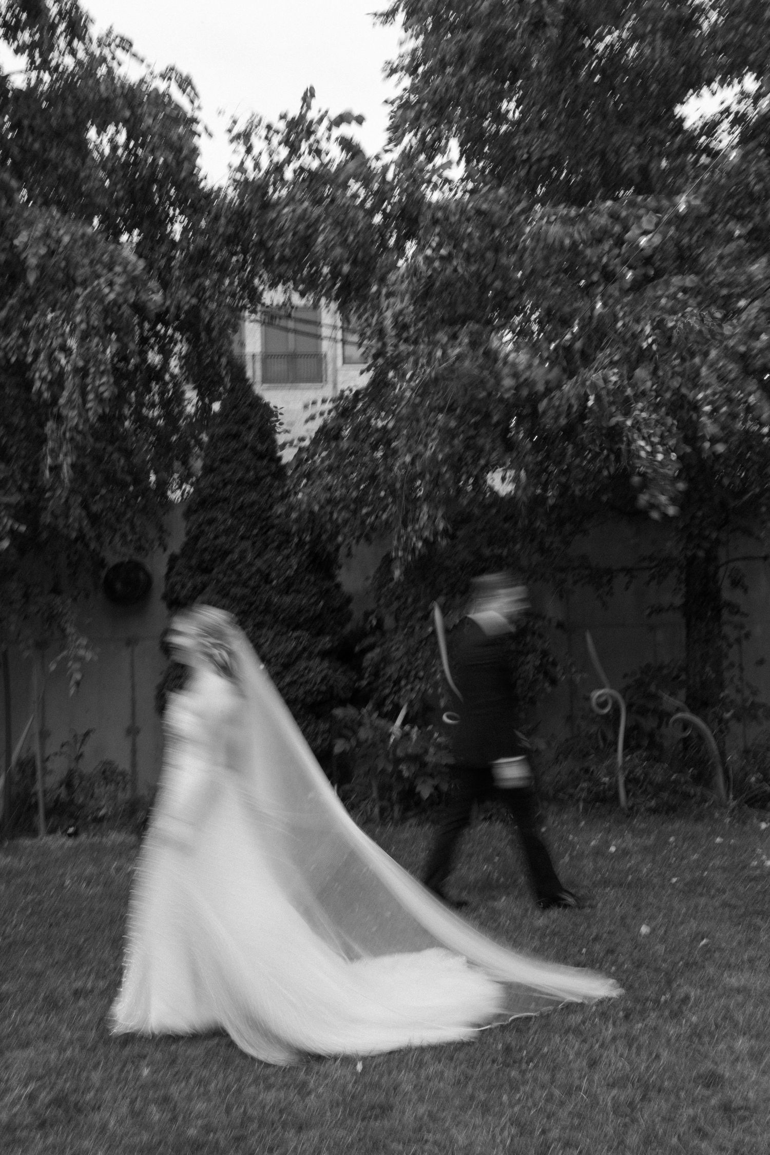 Black and white photo of a bride in a flowing veil and groom walking in a garden, captured with motion blur.
