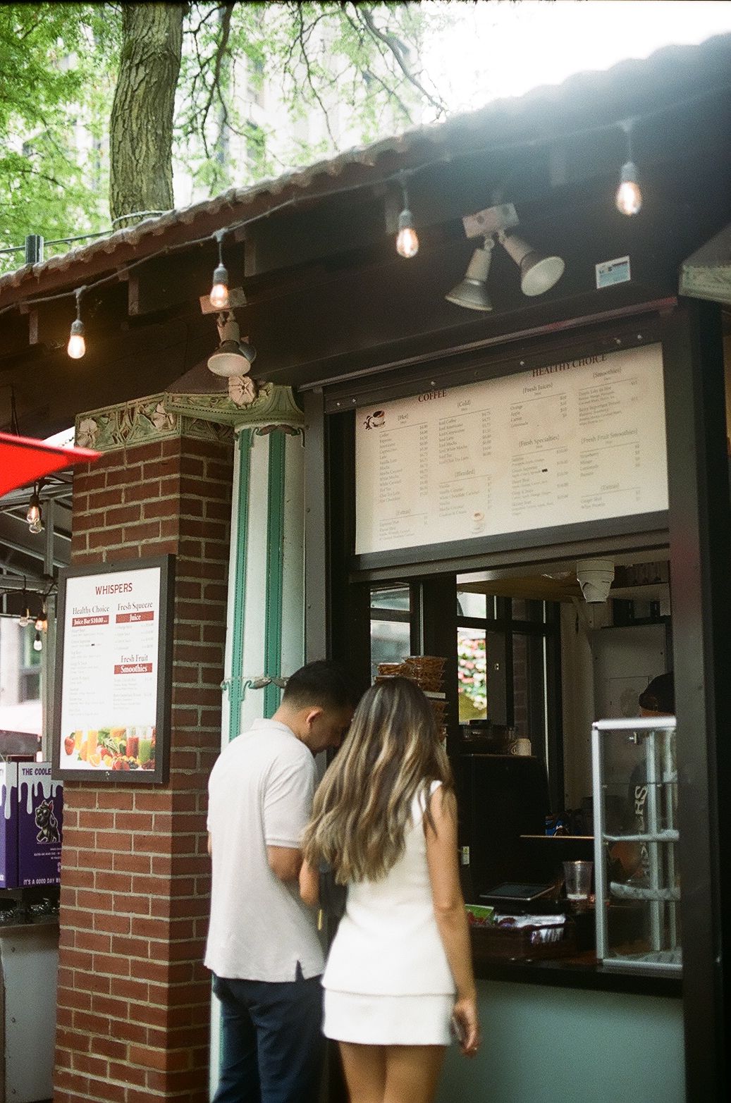 People queue at an outdoor concession stand with string lights and brick walls under a dark overhang.