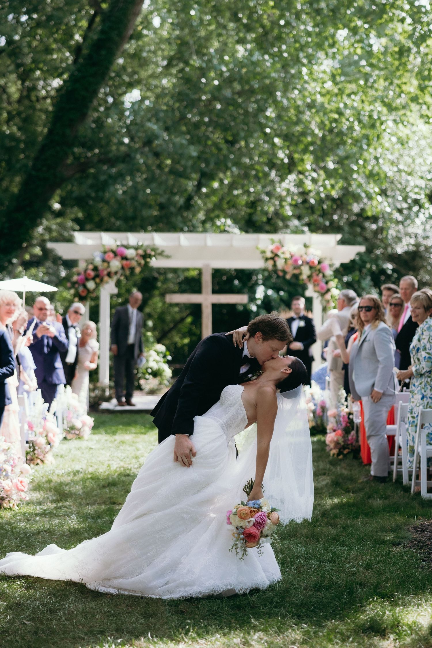 Groom dips bride for a romantic kiss at the altar during outdoor wedding ceremony with floral arch backdrop.