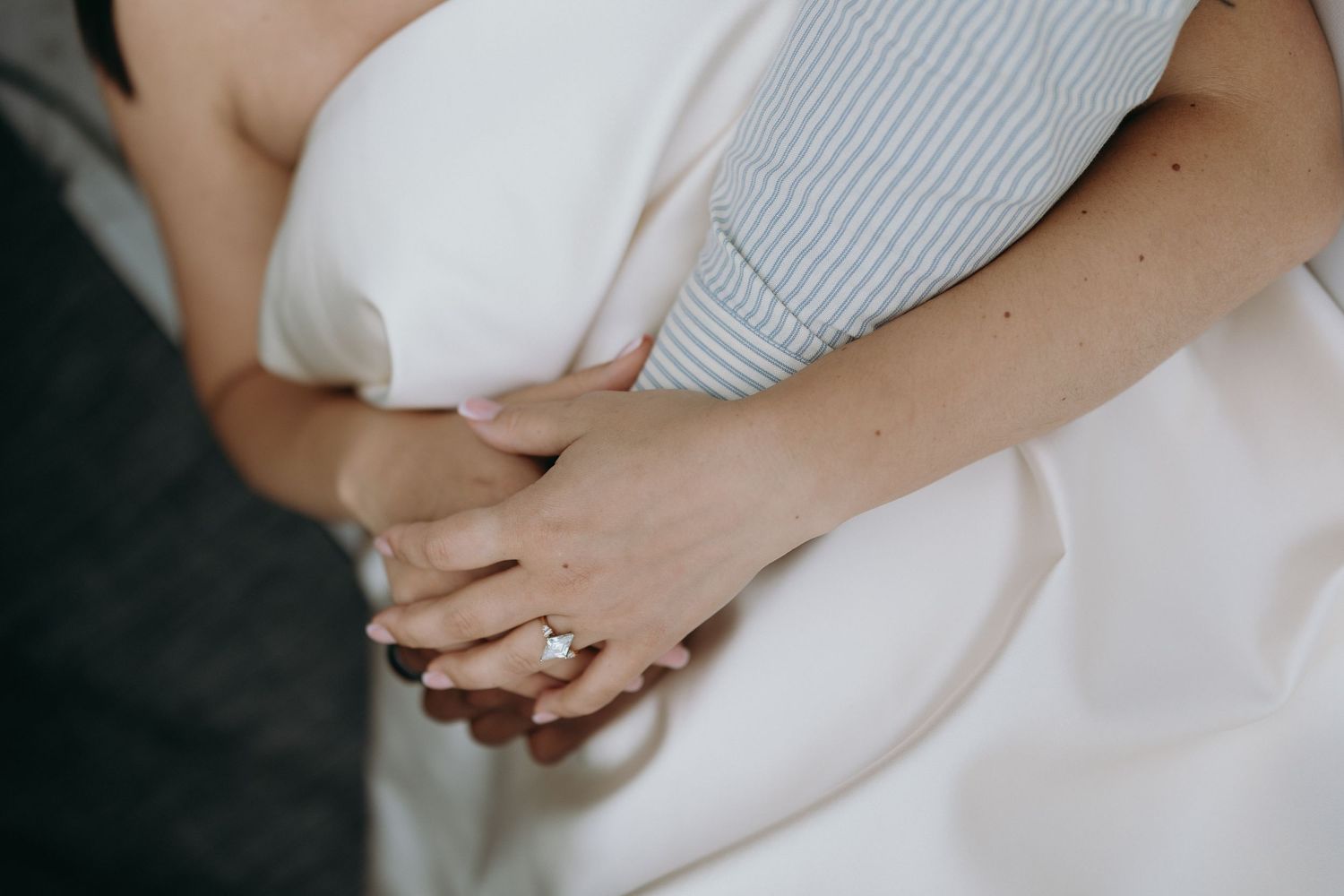 Close-up of a bride's hand with a diamond engagement ring resting on a white wedding dress.