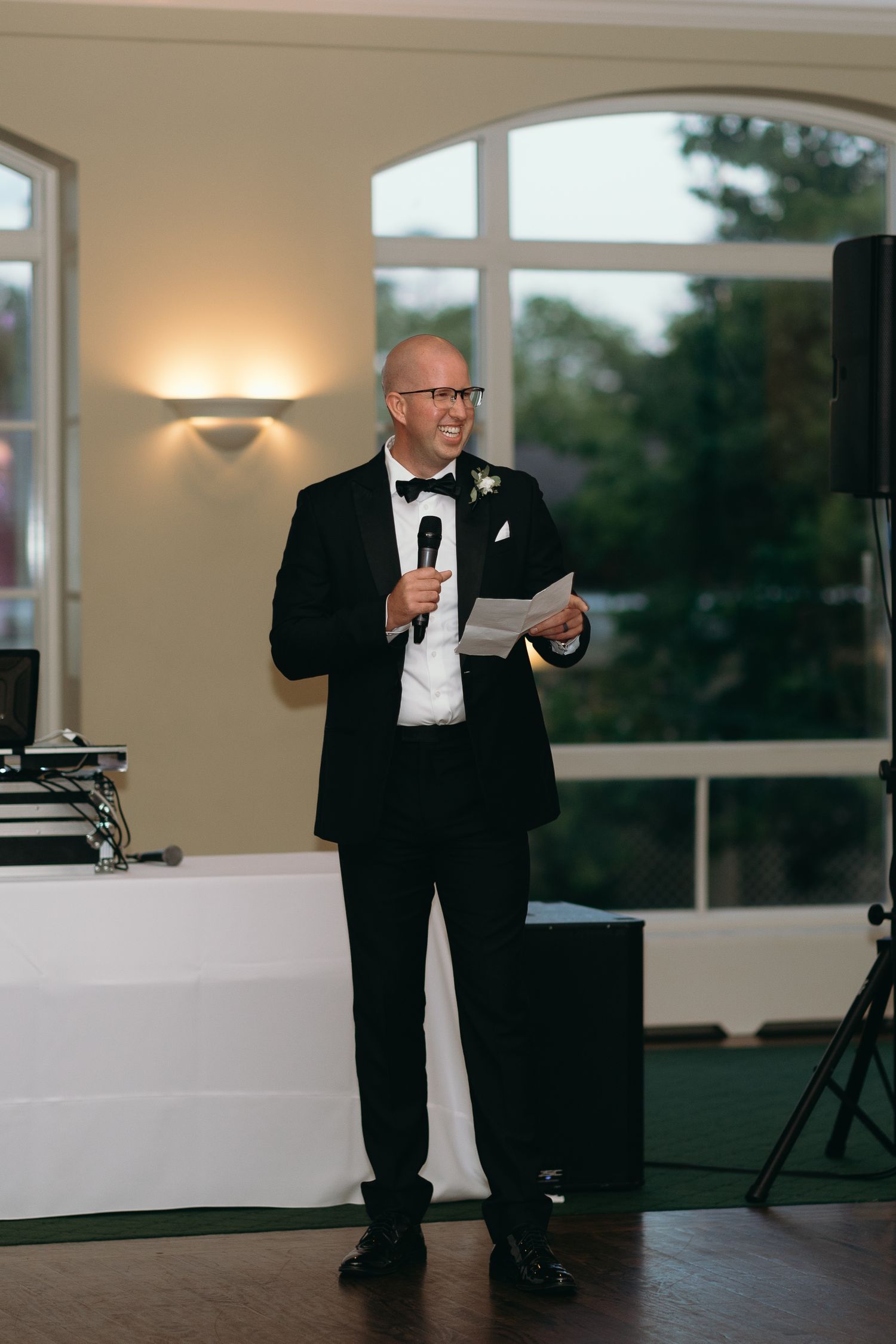 Man in tuxedo delivering speech at formal event with scenic windows in background.