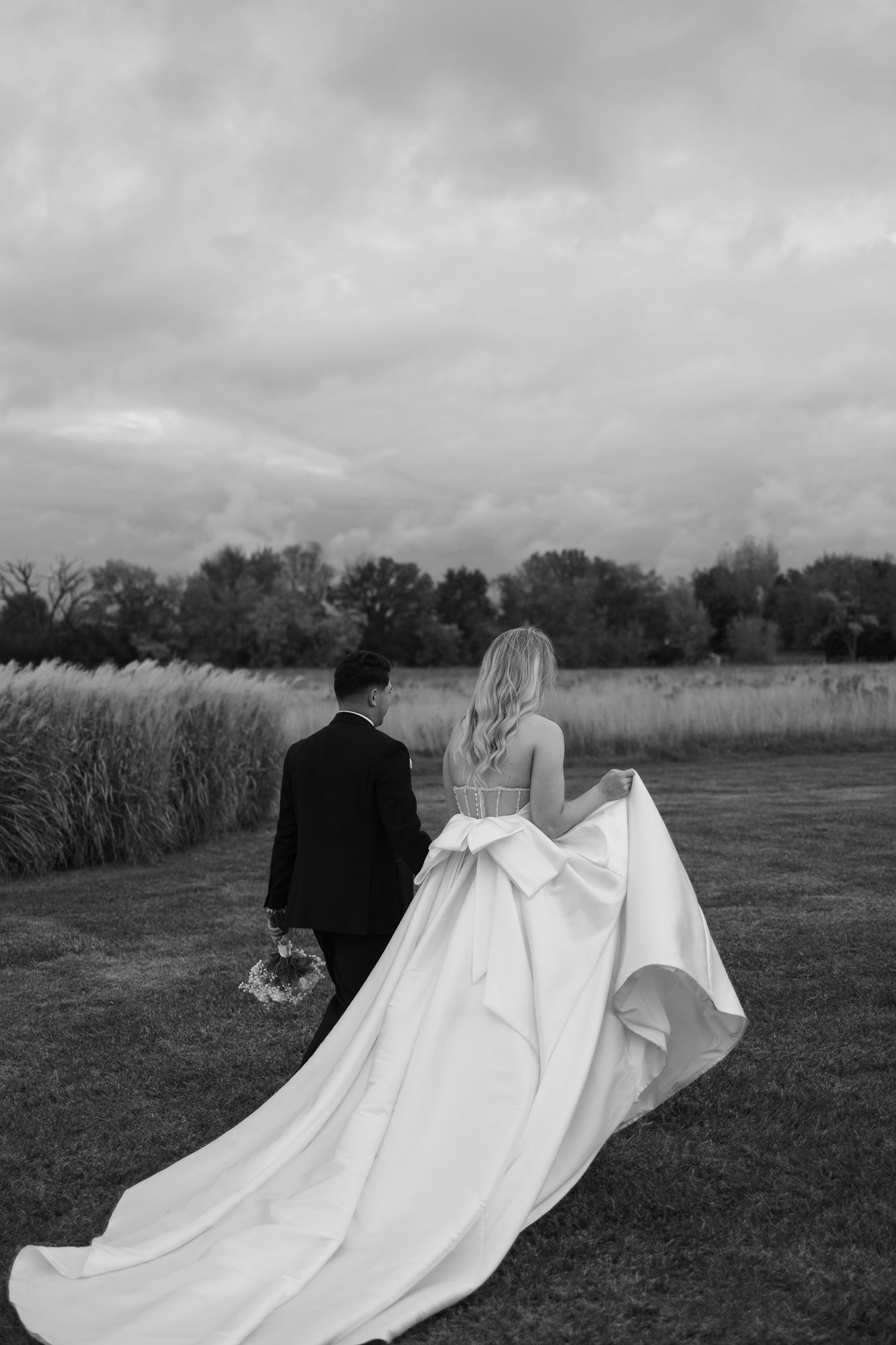 A black and white wedding photo of a couple standing in a field under cloudy skies.
