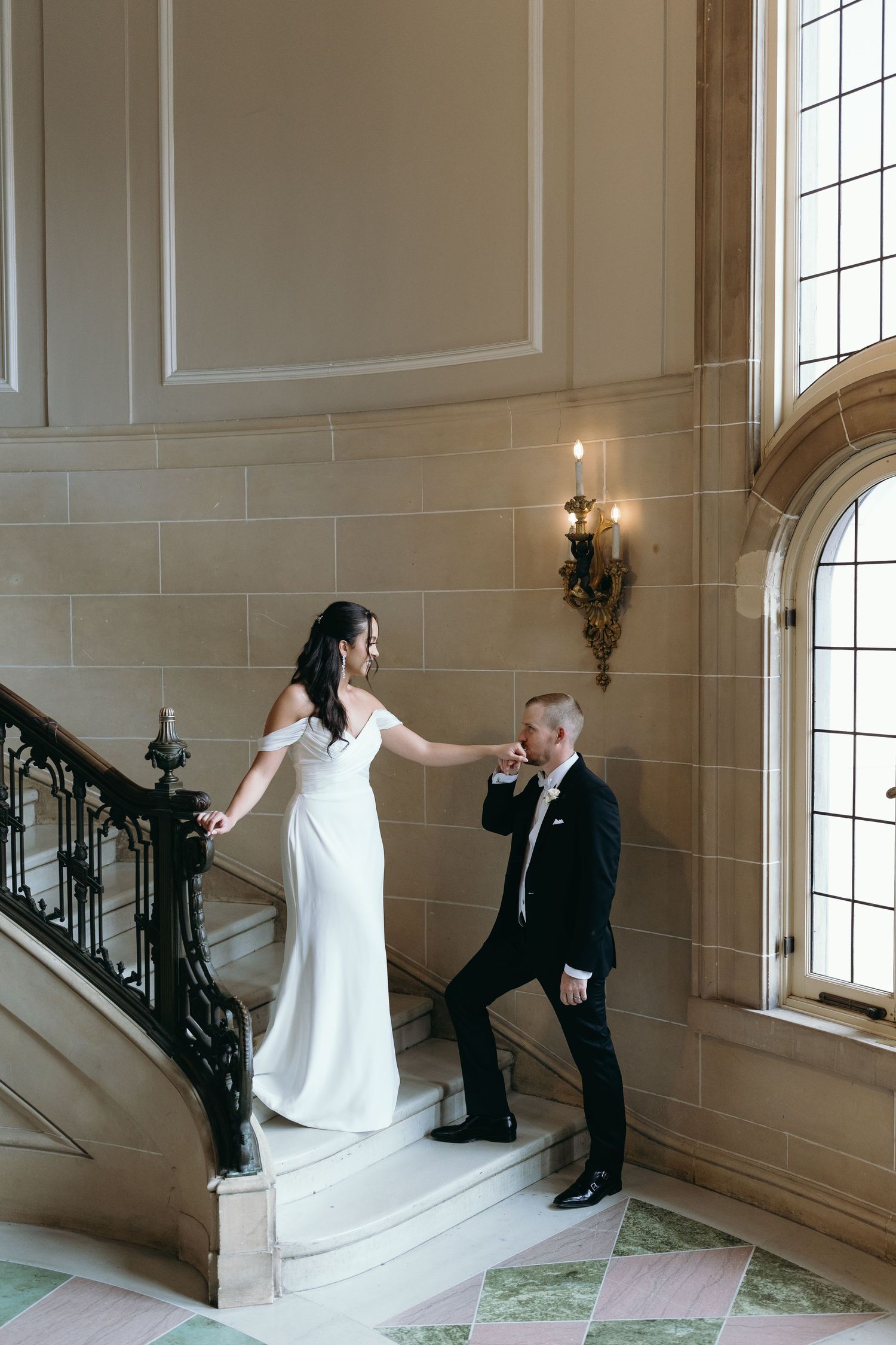 A wedding couple poses on an elegant staircase with arched windows and ornate details.
