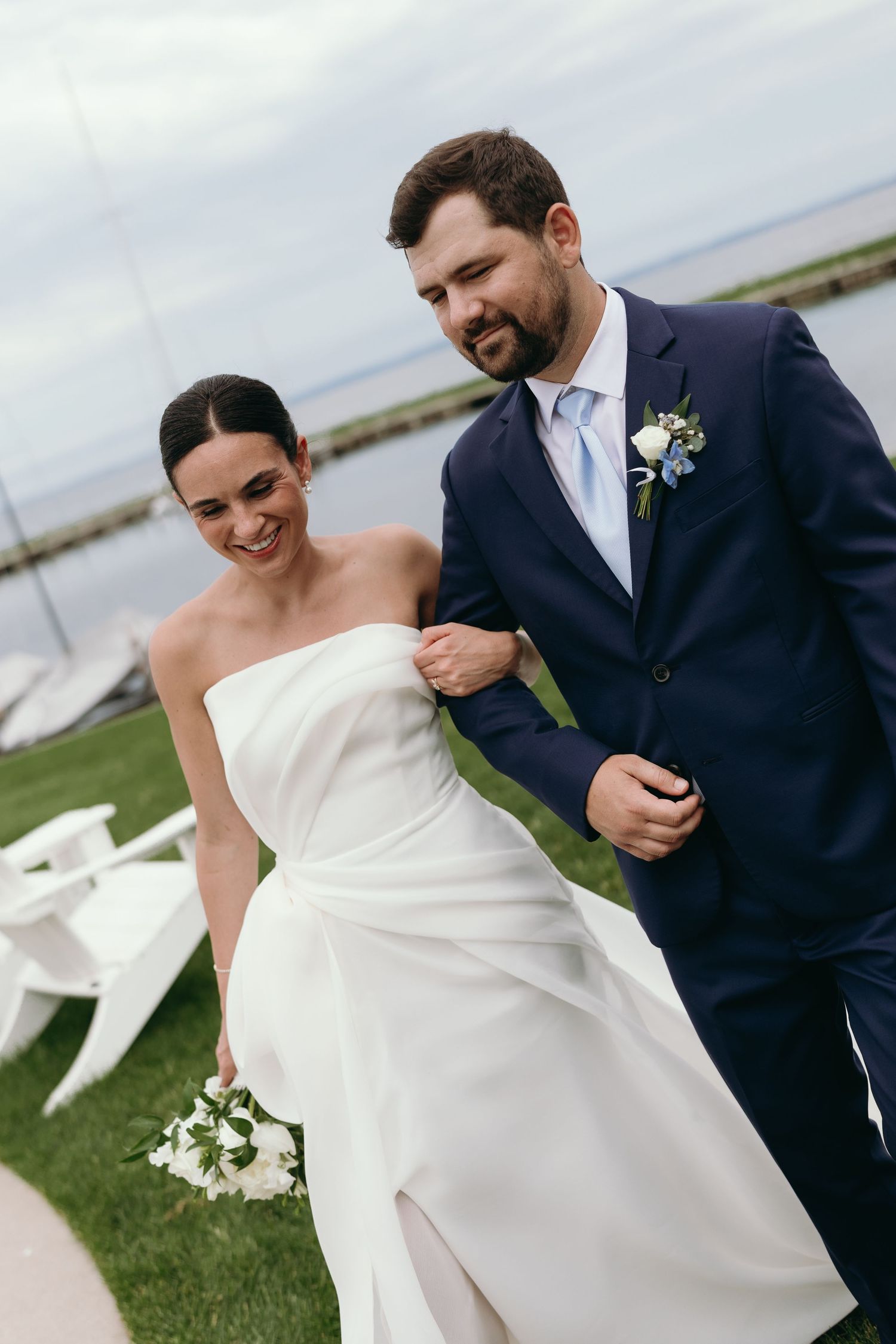 A bride in a strapless white gown and groom in navy suit share a candid moment together outdoors by the water.