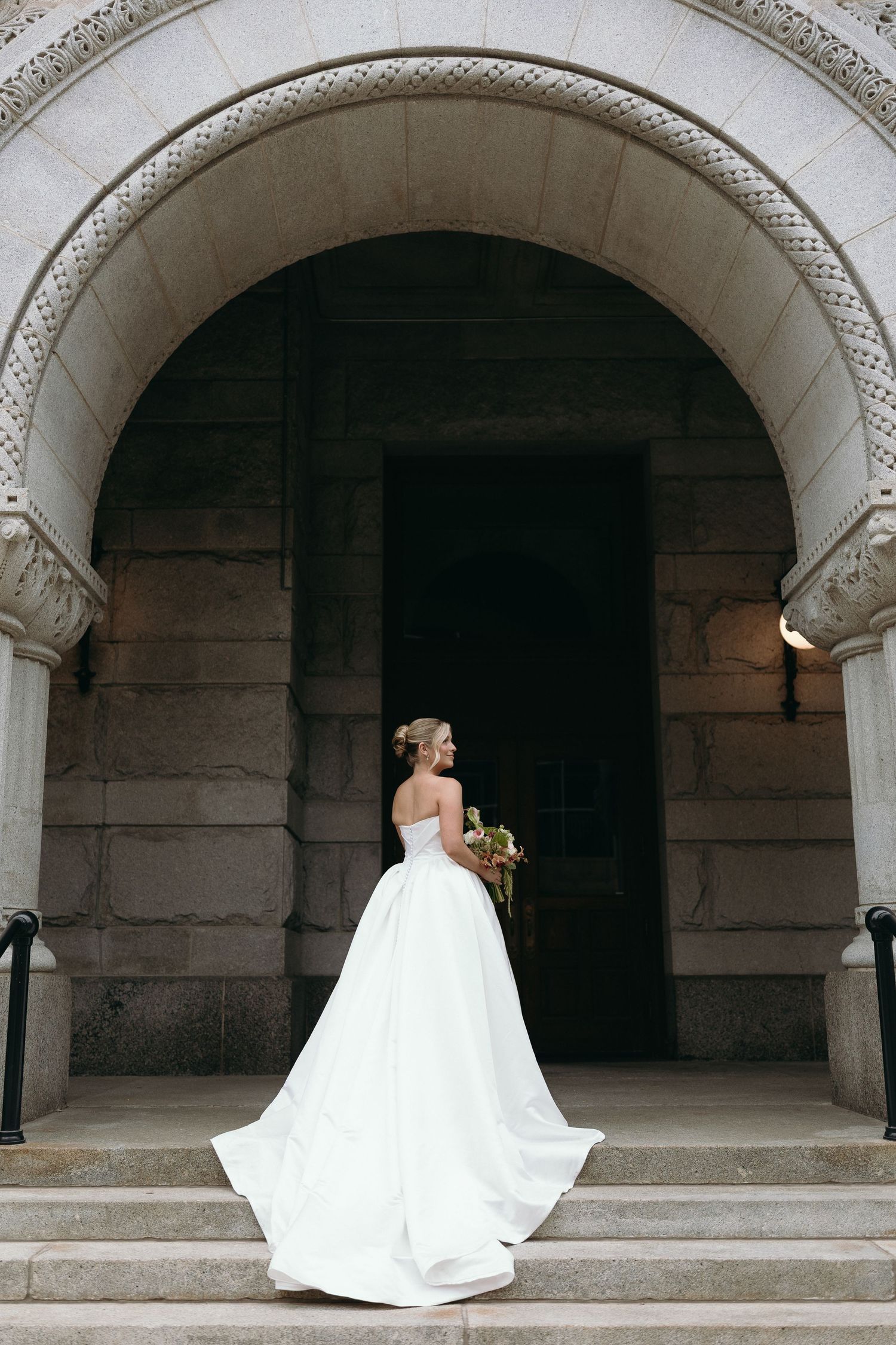 A bride in a flowing white wedding gown poses under a series of stone archways in a historic building.